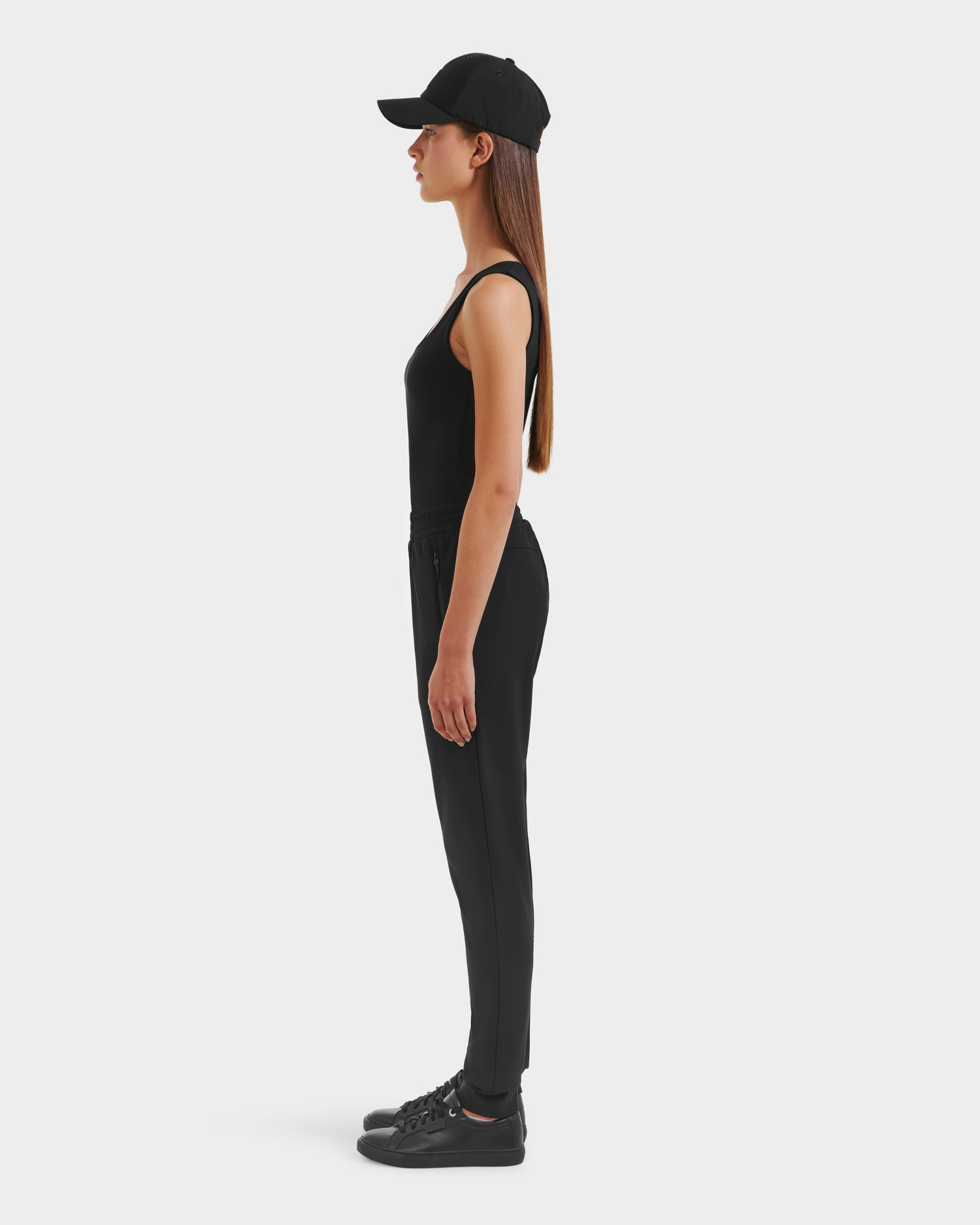 A woman in profile faces left, wearing Rudsak’s DAMERIS - 8525575 Black travel pants with a black cap, sleeveless black top, and black sneakers, set against a plain white background.