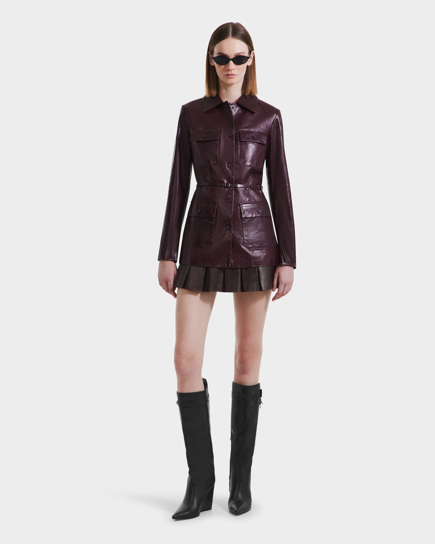 A woman models the Rudsak DANICA - 8125025 Bordeaux tailored leather jacket with front pockets, a matching skirt, black knee-high boots, and black sunglasses against a plain white background.
