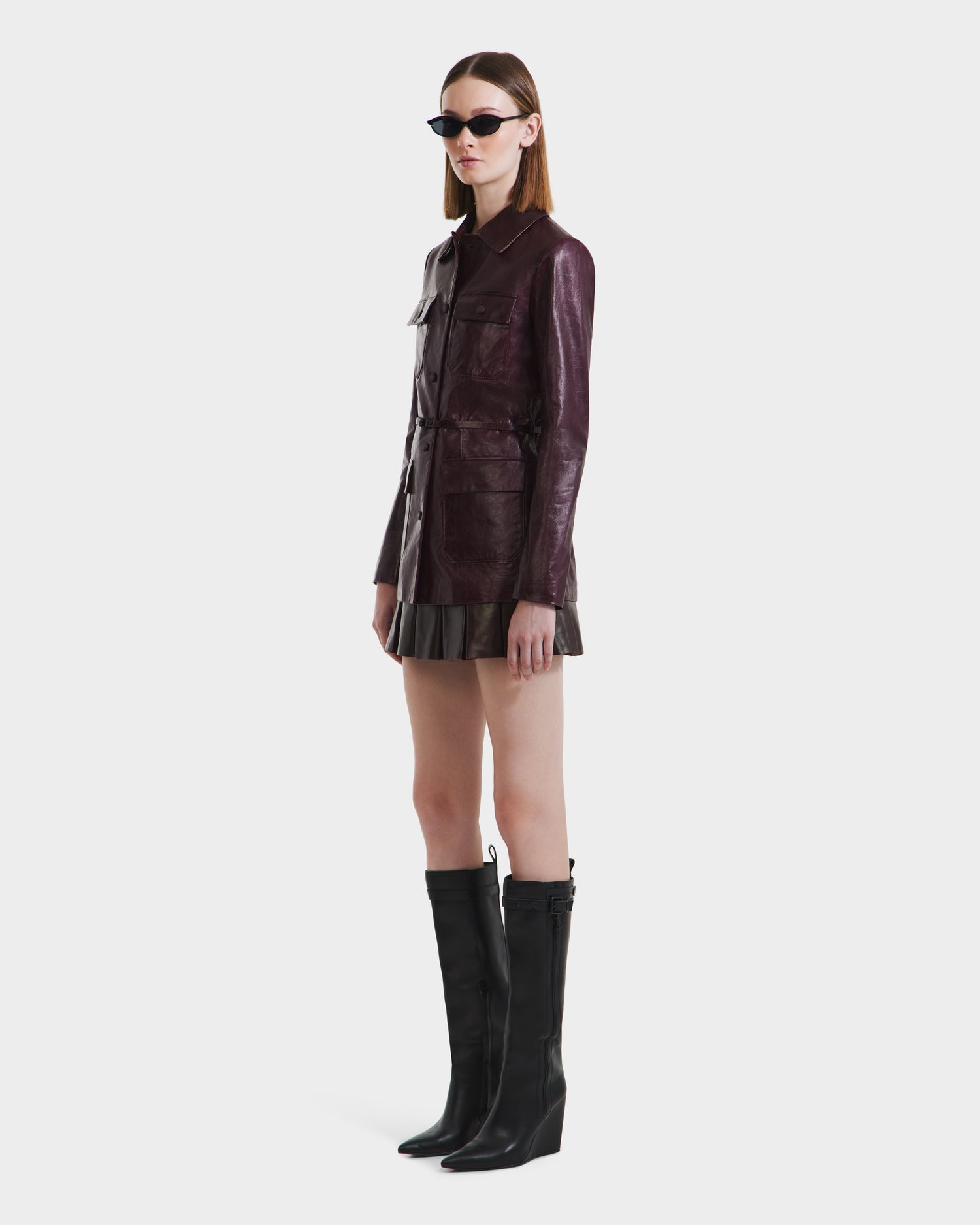 A woman in sunglasses wears the Rudsak DANICA - 8125025 Bordeaux womens tailored leather jacket, paired with a short skirt and knee-high black boots, standing against a plain white background.