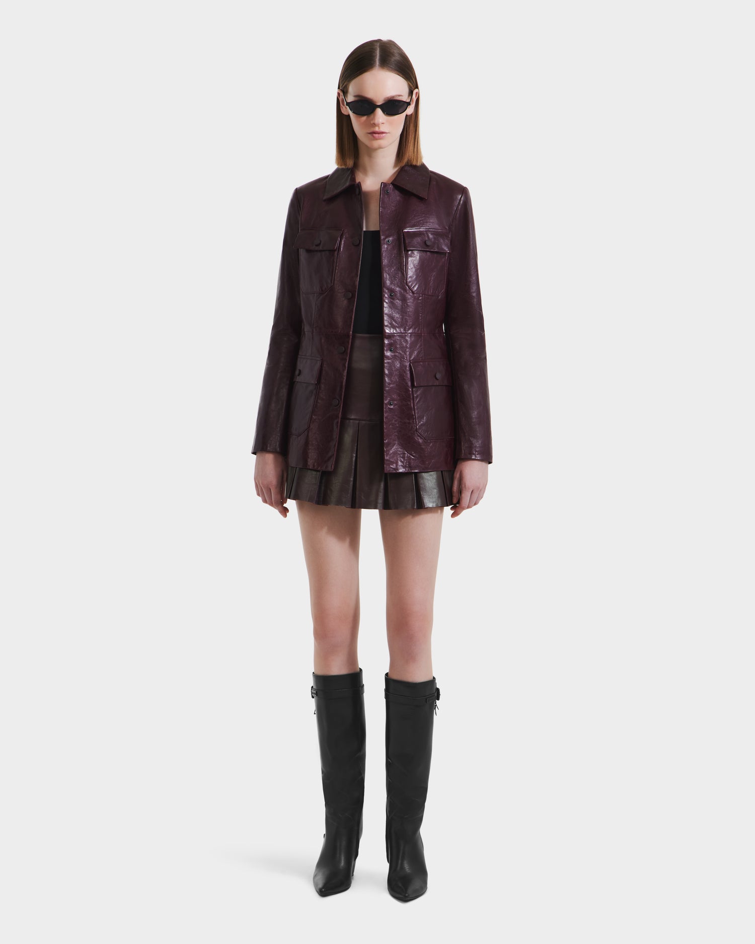 A woman in dark sunglasses and knee-high black boots wears the Rudsak DANICA - 8125025 Bordeaux glossy burgundy leather jacket with a tailored fit and front pockets, paired with a short pleated skirt, against a plain white background.