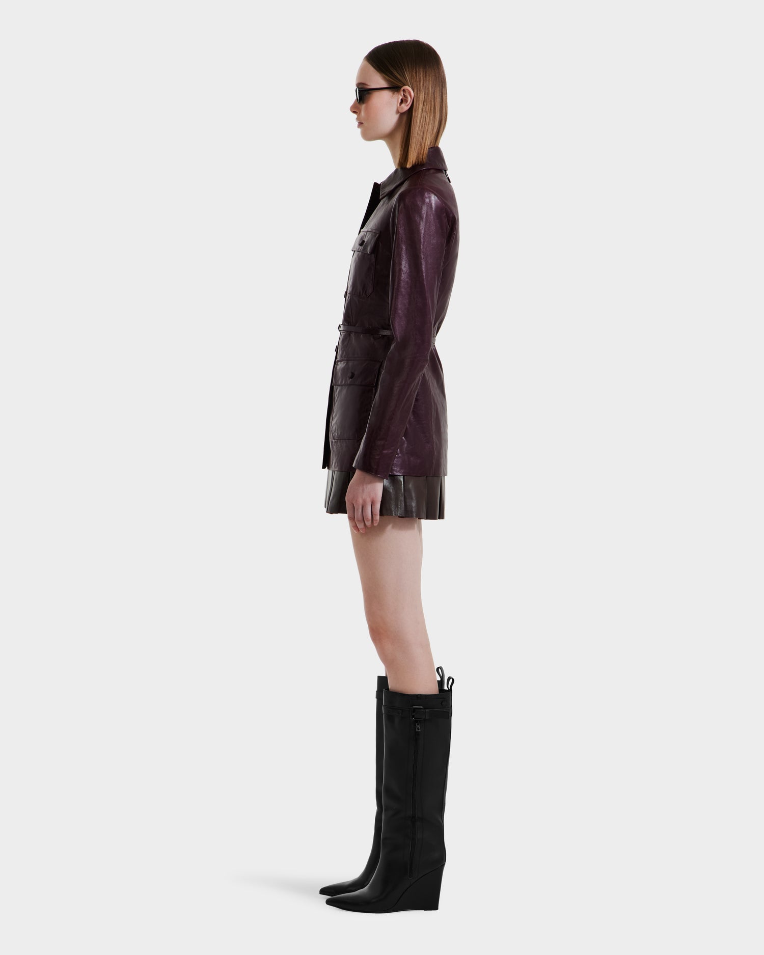 A woman in profile wears Rudsak’s DANICA - 8125025 Bordeaux leather jacket, sunglasses, a short skirt, and knee-high black wedge boots against a plain white background.