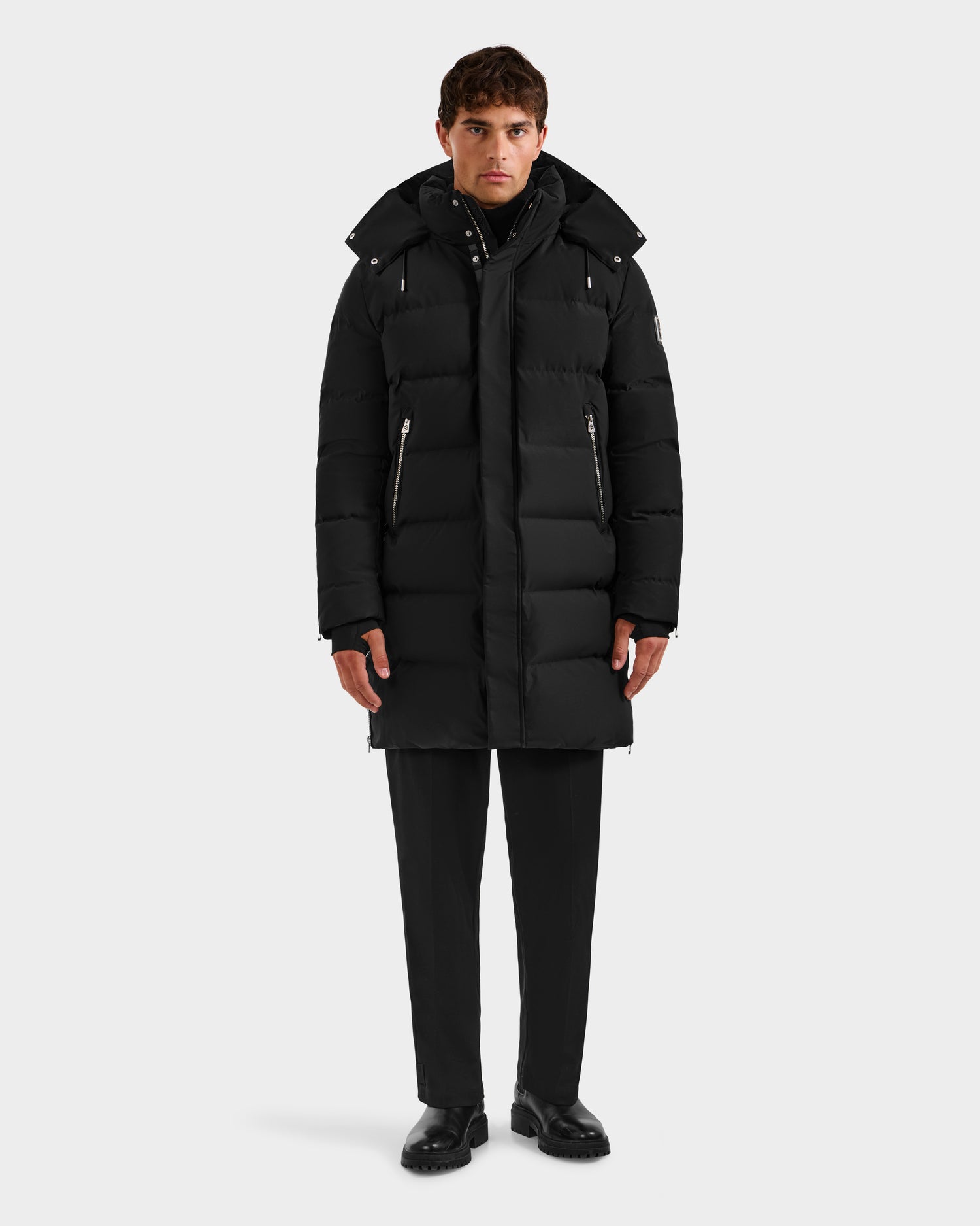 A man stands facing forward in the Rudsak DAO H - 6125583 Black men’s quilted down jacket with a hood, paired with black pants and shoes—stylish winter outerwear—set against a plain light gray background.