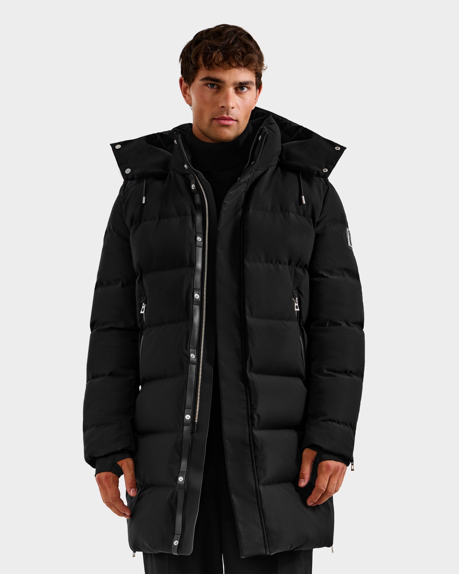 A man stands against a light gray background, wearing the Rudsak DAO H - 6125583 Black men's long quilted down jacket with hood and zippered front—perfect for cold-weather performance and stylish winter outerwear.