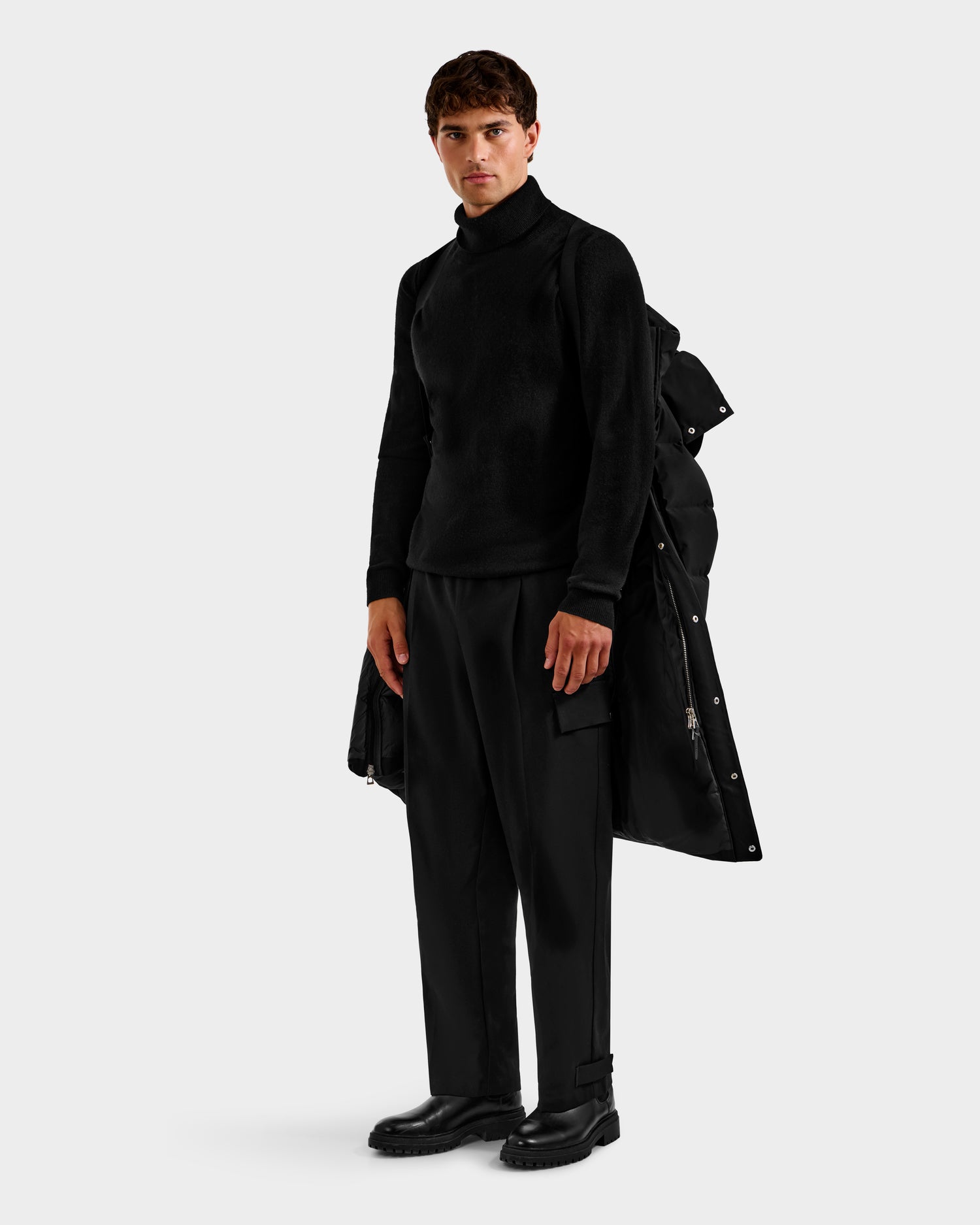 A man stands against a plain background wearing the Rudsak DAO H - 6125583 Black coat draped over his left shoulder, paired with a black turtleneck sweater, wide-leg pants, and boots for cold-weather style.