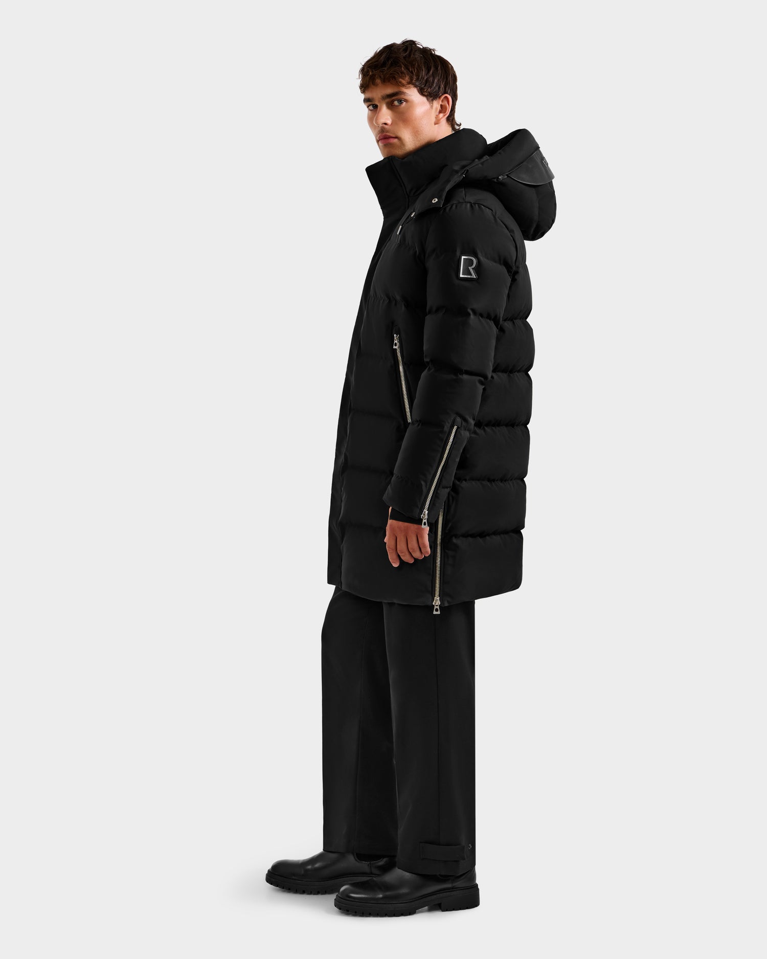 A man in profile wears the Rudsak DAO H - 6125583 Black men’s quilted down jacket with hood. The long puffer has side zippers, a small sleeve logo patch, and is paired with black pants and boots against a plain white background.