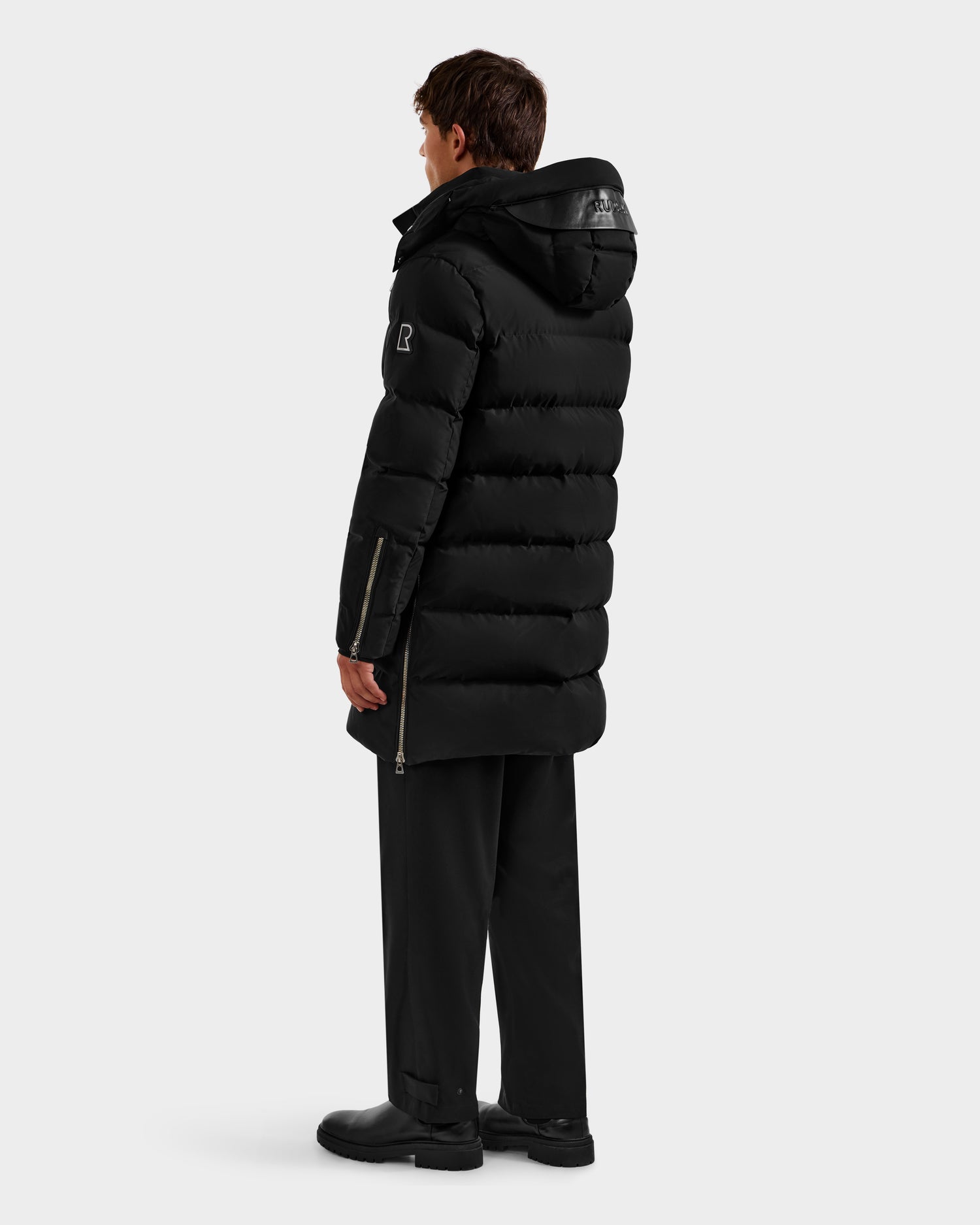 A person stands facing away, wearing the Rudsak DAO H - 6125583 Black men’s quilted down jacket with a hood, black pants, and black shoes against a plain light gray background—styled for premium cold-weather performance.