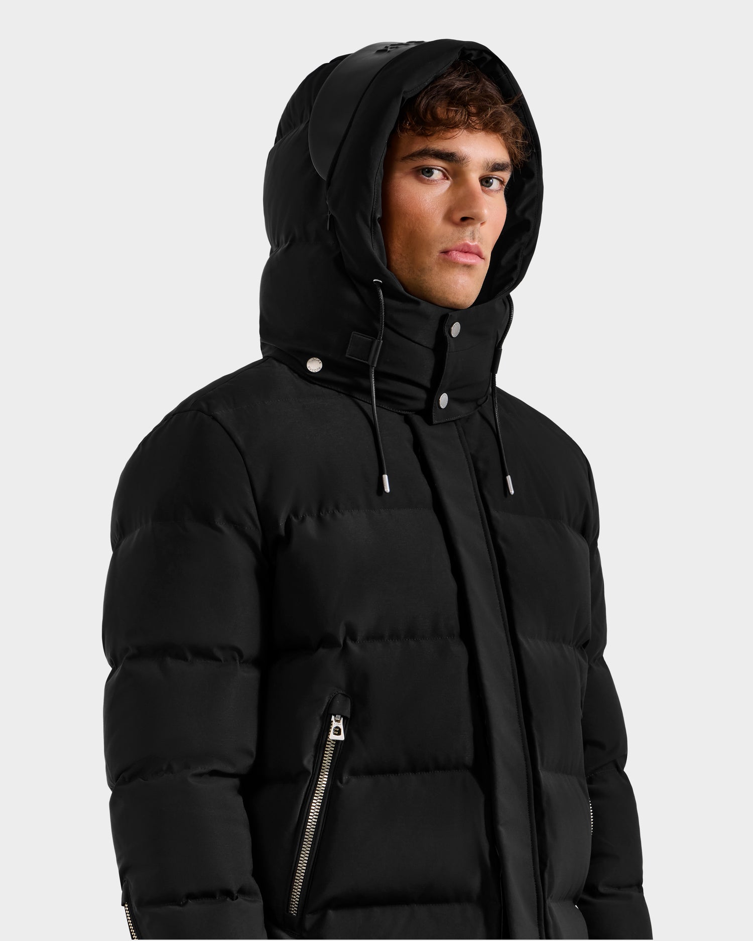 A young man models the Rudsak DAO H - 6125583 Black men's quilted down jacket with hood and silver zippers, highlighting essential winter outerwear against a plain light gray background.