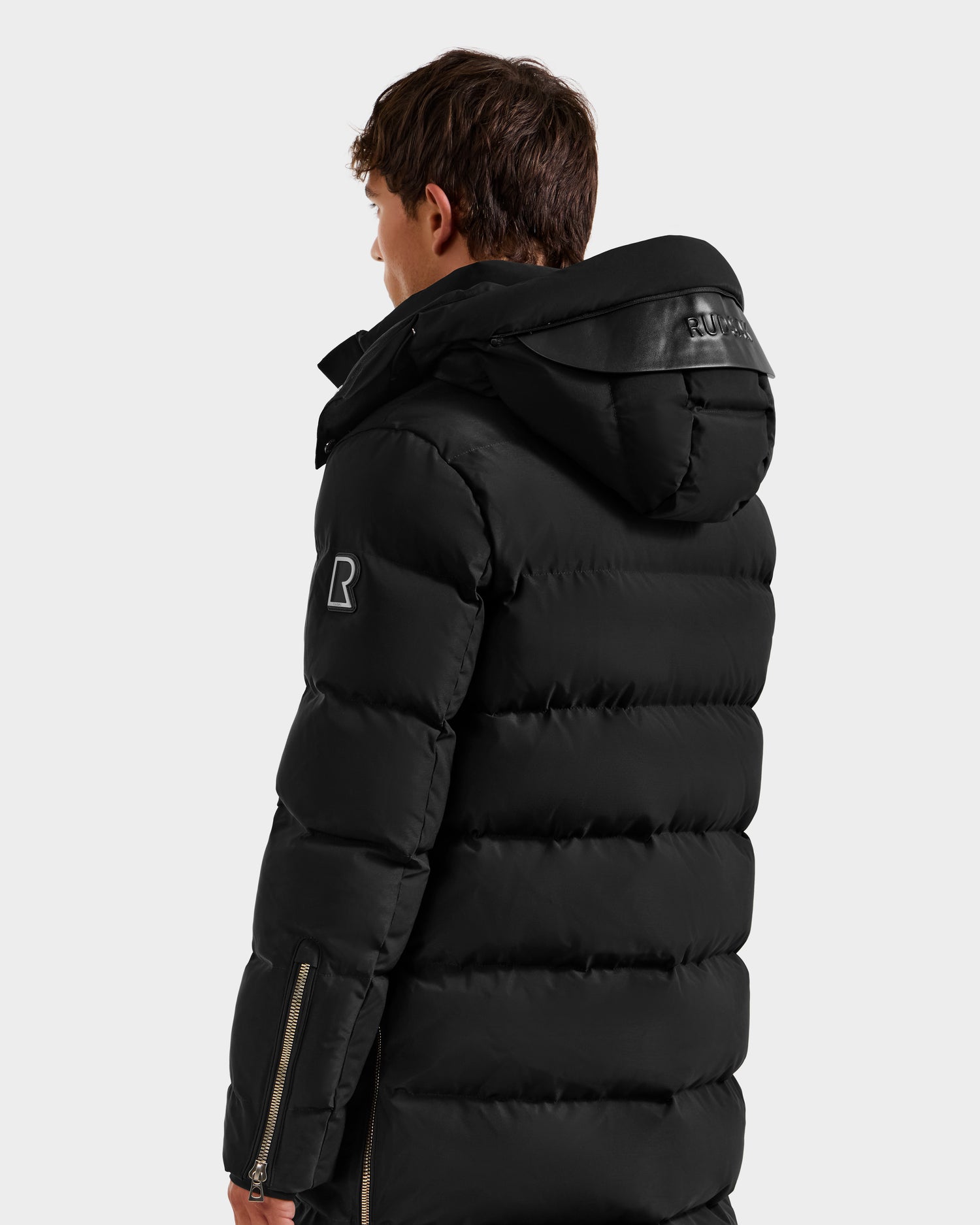 A person with short brown hair wears the Rudsak DAO H - 6125583 Black men’s quilted down jacket, featuring a hood and sleeve zipper detail, standing against a light gray background to showcase winter outerwear.