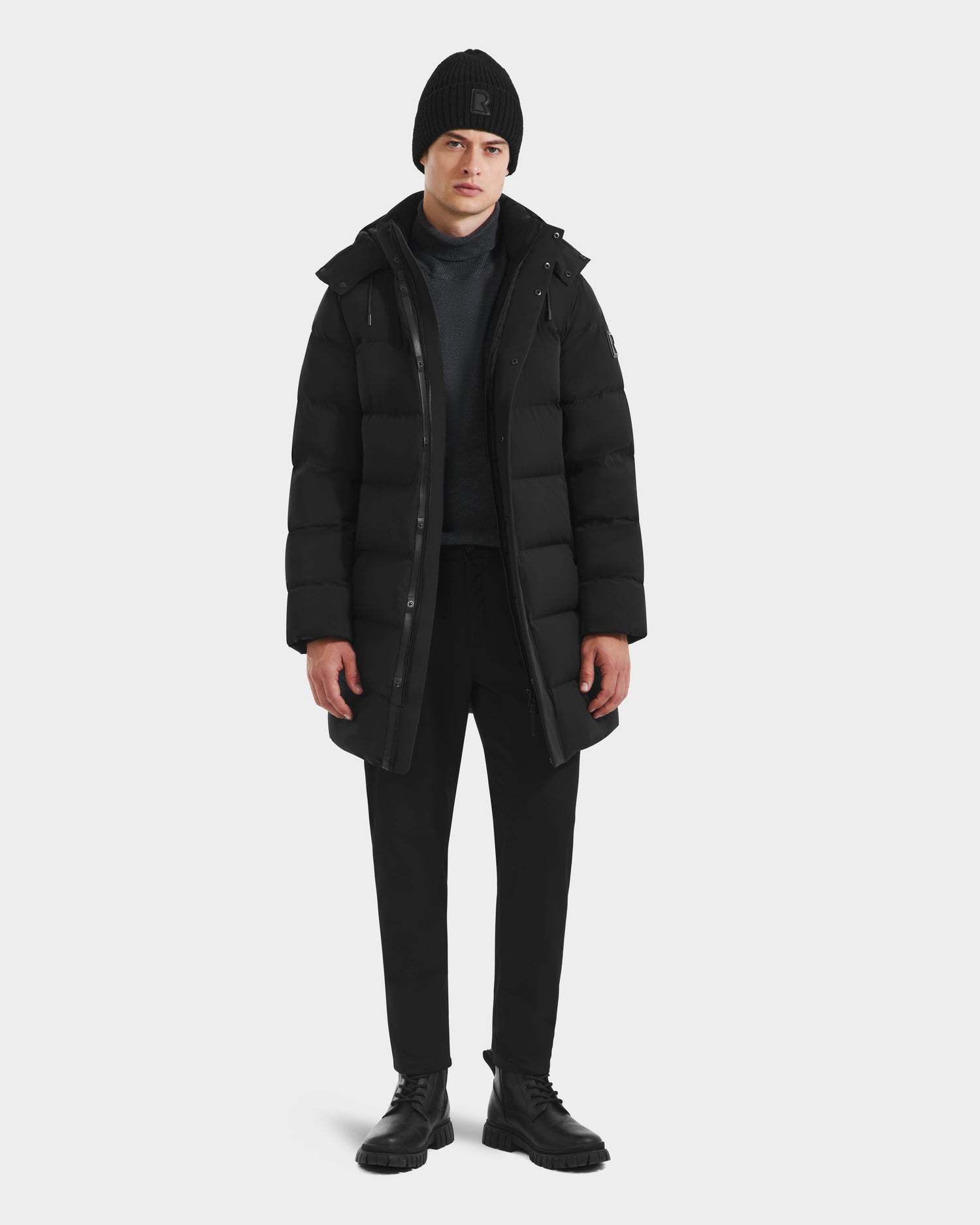 A person faces forward against a plain white background, wearing the Rudsak DAO - 6124604 Black mens puffer jacket, paired with a black beanie, dark sweater, black pants, and black boots.