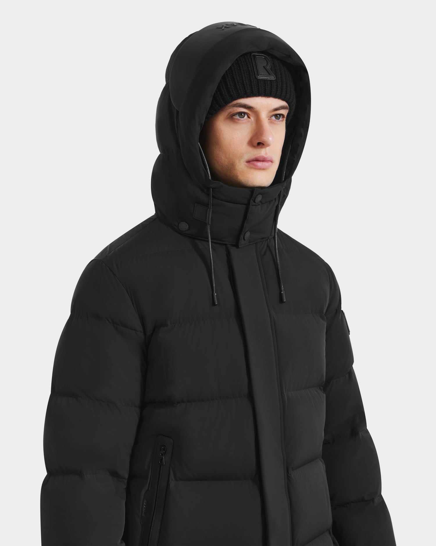 A person wearing the Rudsak DAO - 6124604 Black mens puffer jacket with the hood up and a black beanie, looking slightly to the side against a plain light background.