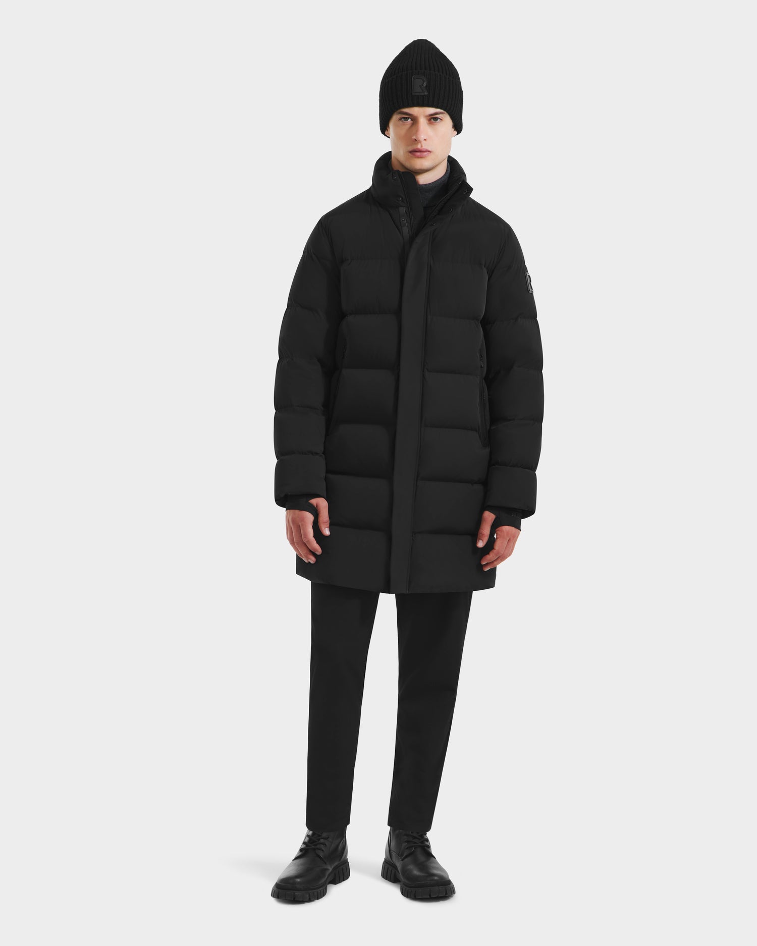 A person stands facing forward in a Rudsak DAO - 6124604 Black men’s puffer jacket, black beanie, black pants, and black shoes against a plain white background.
