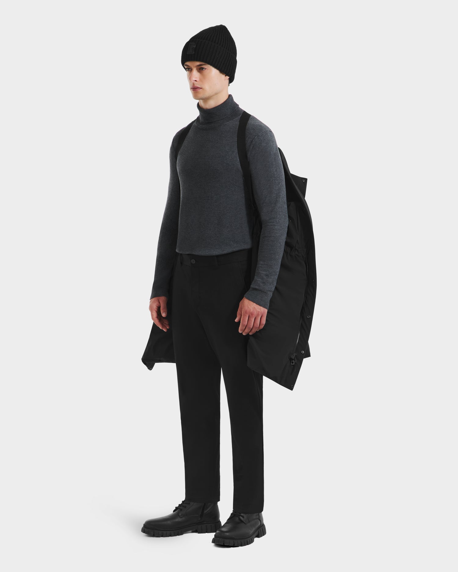 A person stands against a plain white background, holding the weather-resistant Rudsak DAO - 6124604 Black down coat over one shoulder, dressed in a black beanie, gray turtleneck sweater, black pants, and black shoes.