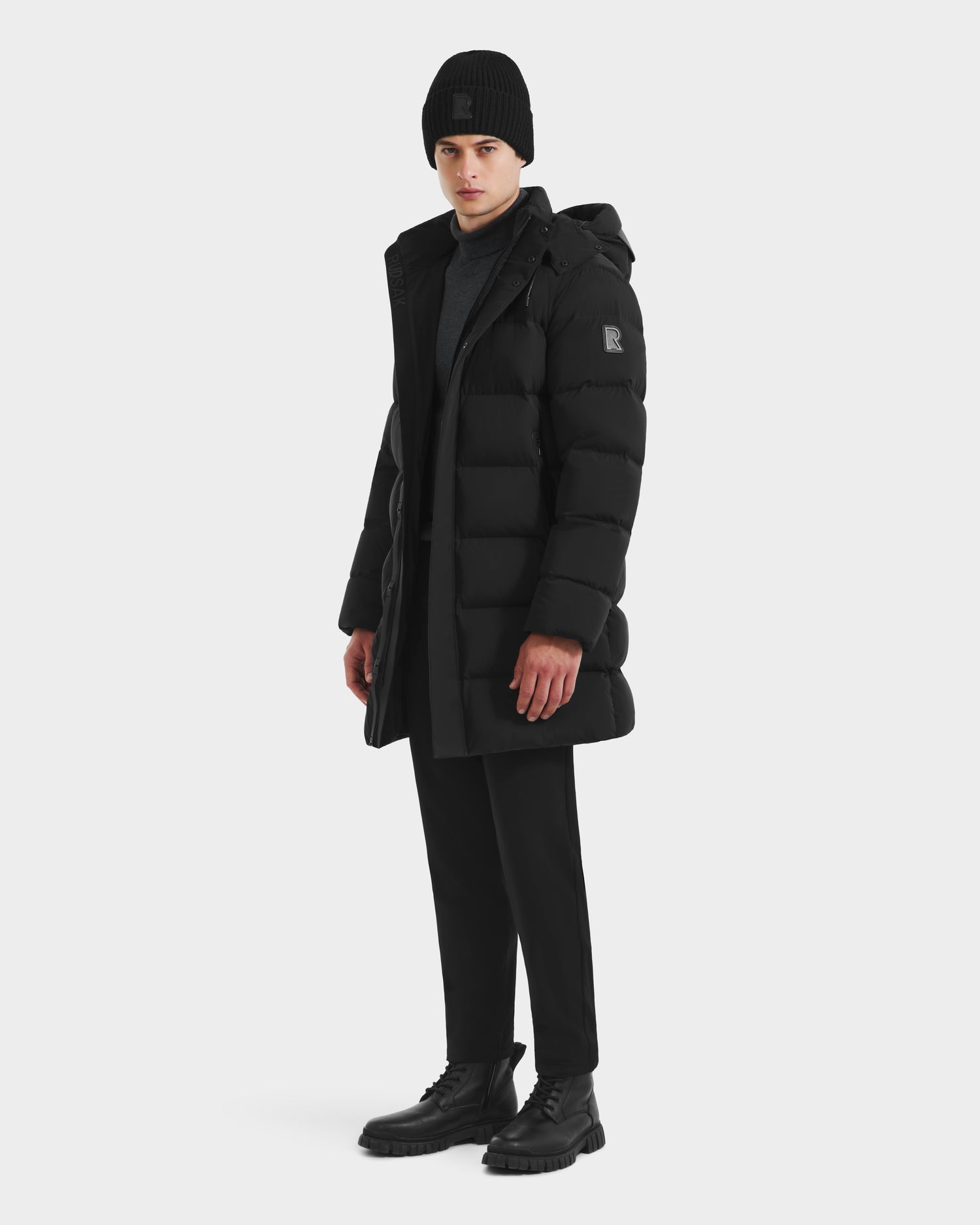 A man models the Rudsak DAO - 6124604 Black mens puffer jacket with black pants, lace-up boots, a beanie, and a dark turtleneck sweater, standing against a plain white background.