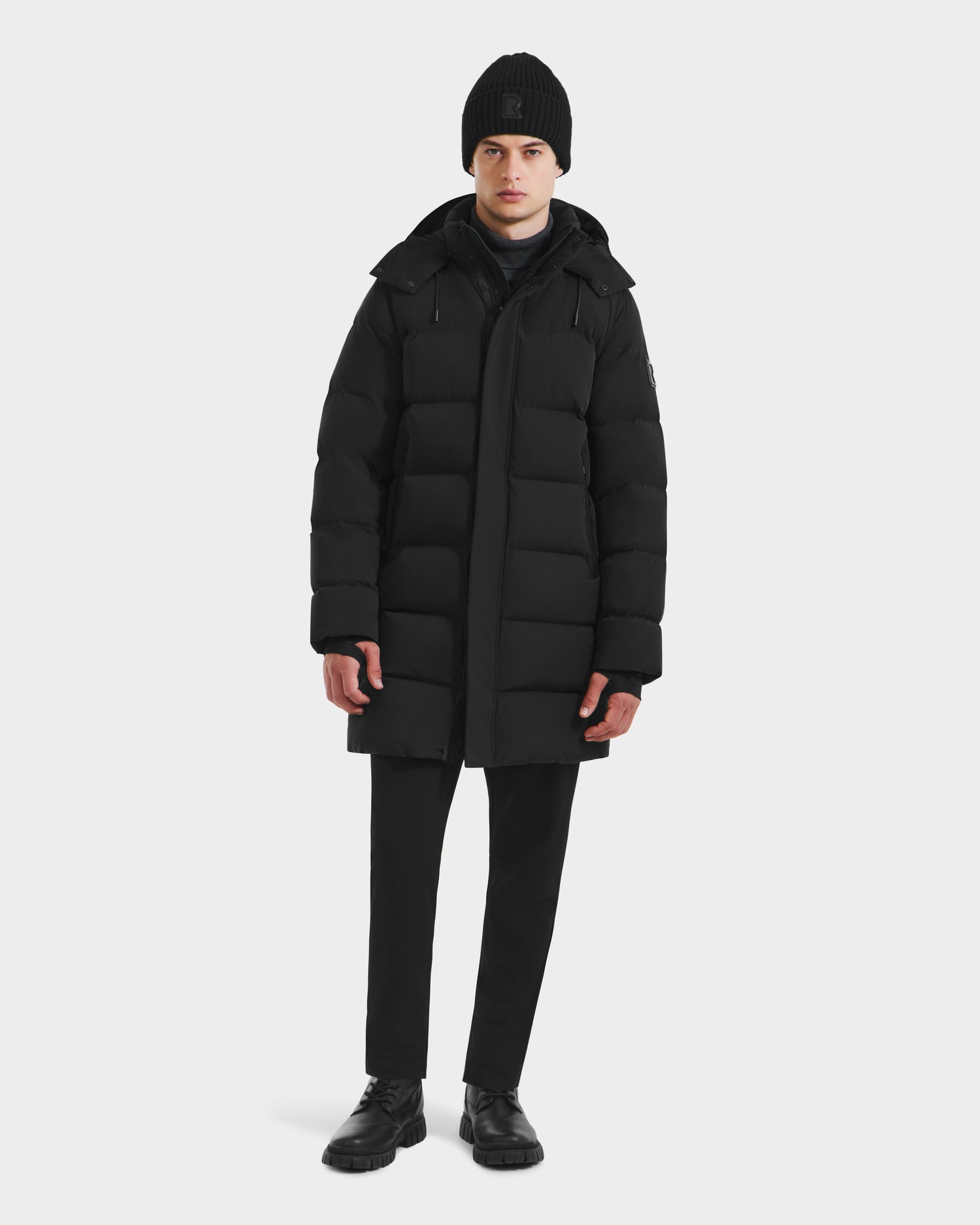 A person wears the Rudsak DAO - 6124604 Black weather-resistant down coat, a black knit beanie, black pants, and black shoes while facing forward against a plain white background.