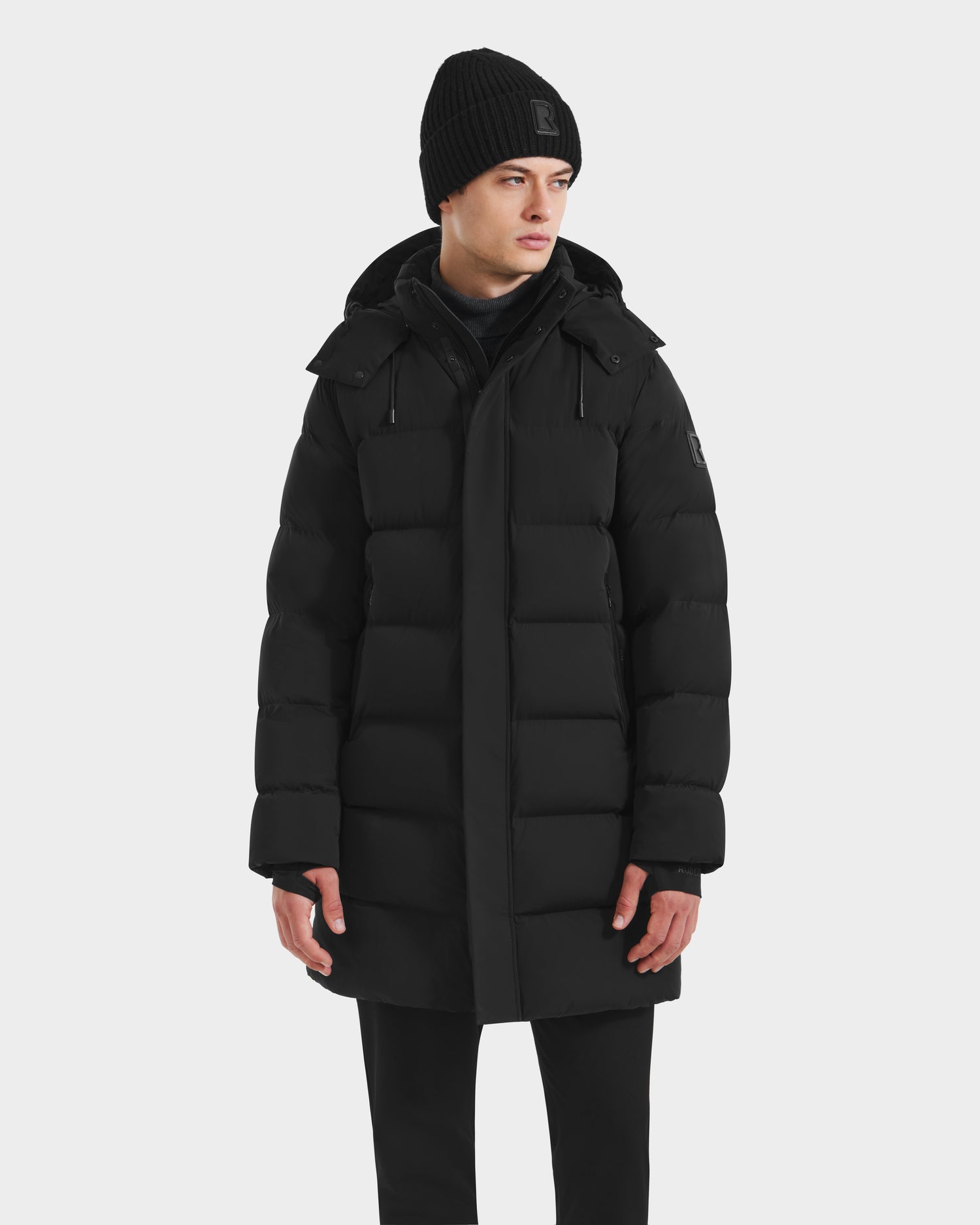 A person wears the Rudsak DAO - 6124604 Black mens puffer jacket, black knit beanie, and black pants while standing against a plain white background and looking slightly to the side.