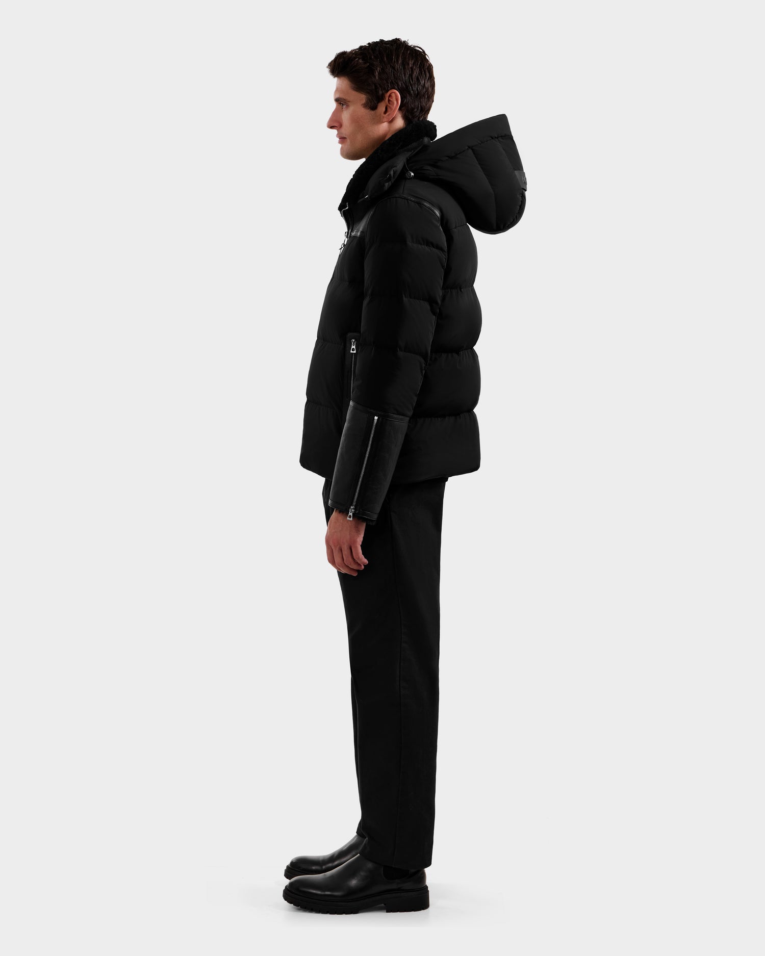 A man stands in profile wearing the Rudsak DAVID - M125508 Black down puffer jacket, paired with black pants and boots, set against a plain light gray background.