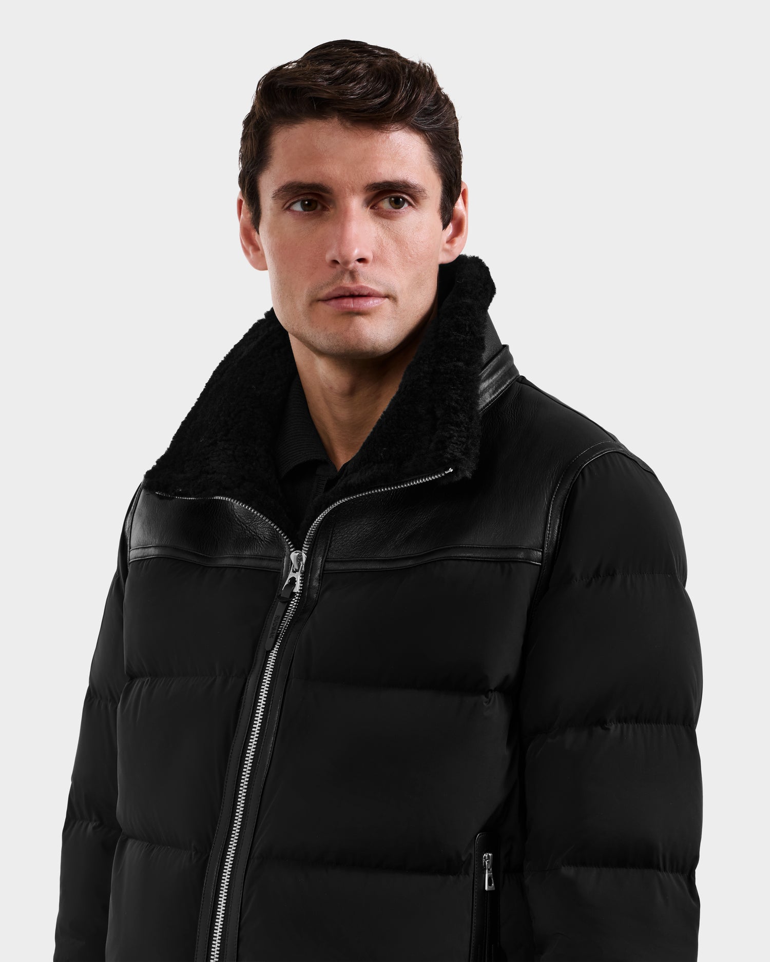 A man with short dark hair wears the Rudsak DAVID - M125508 Black men’s shearling puffer jacket with a faux fur collar, looking slightly to the side against a plain light background.