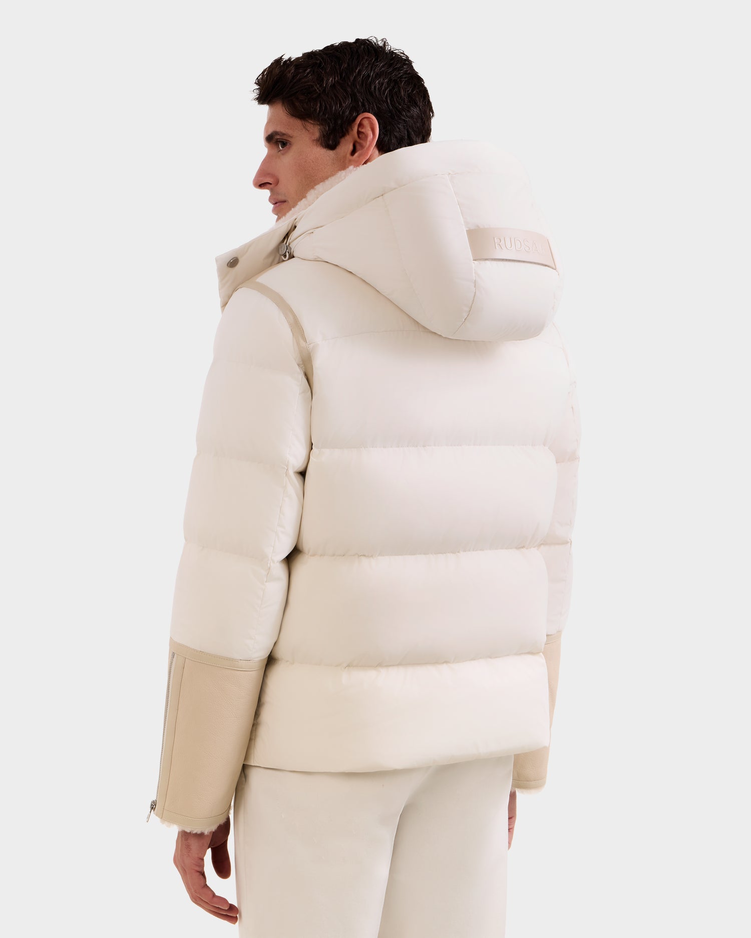 A person stands facing away, showcasing the Rudsak DAVID - M125508 Vanilla: a white winter-ready men’s shearling down puffer with beige accents, hood, high collar, and sleeve zipper details.