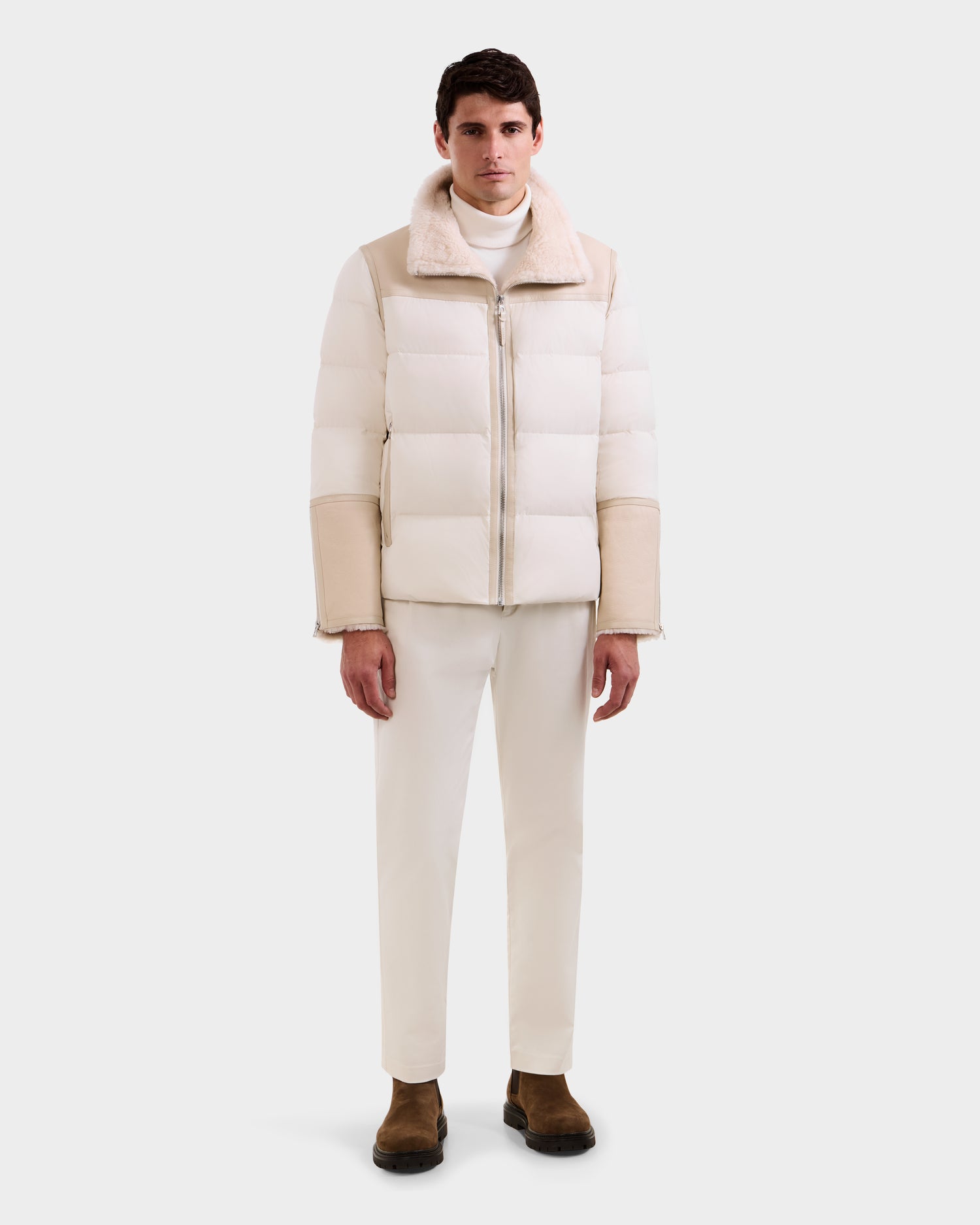 A man stands against a plain white background wearing the Rudsak DAVID - M125508 Vanilla men’s shearling down puffer jacket, paired with a white turtleneck, pants, and brown shoes. He looks straight ahead with his hands by his sides.