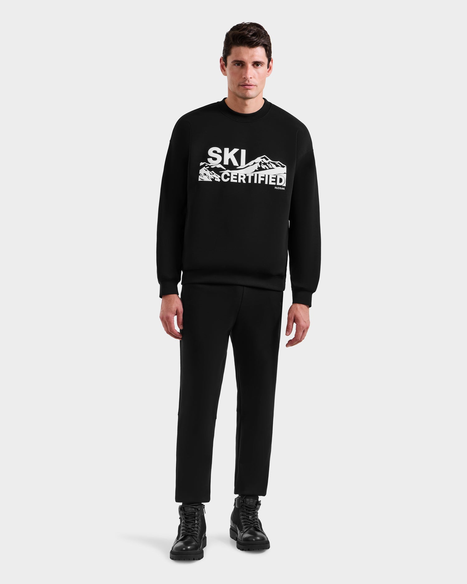 A man faces forward in the Rudsak DECLAN - 6525648 Black men’s crewneck sweatshirt with SKI CERTIFIED and mountain graphics, styled with black pants and boots against a plain light background.