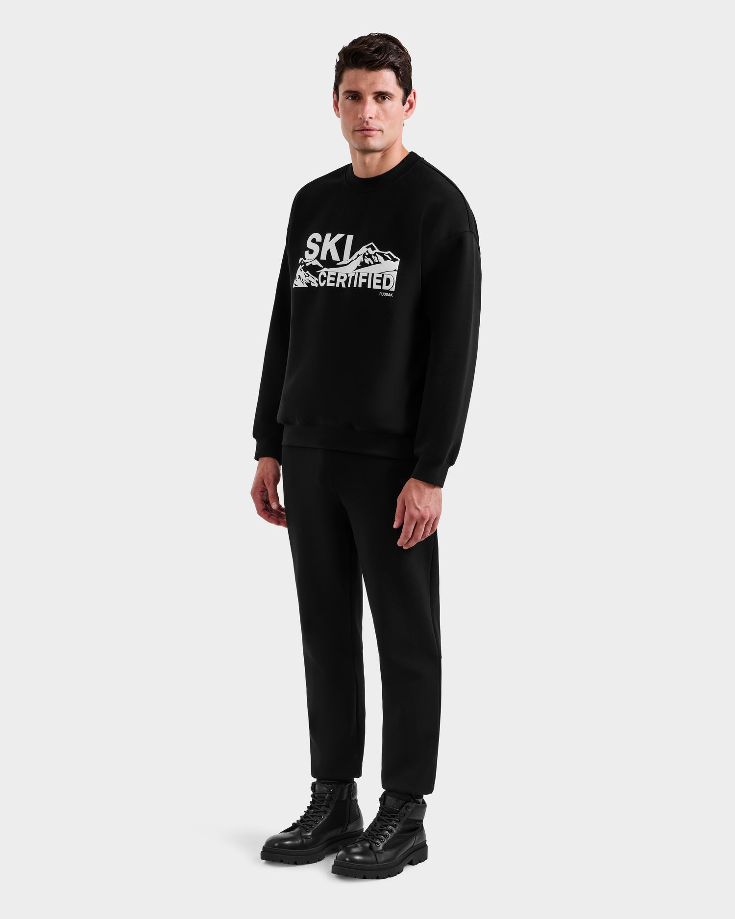 A man faces forward against a plain background wearing the Rudsak DECLAN - 6525648 Black men’s crewneck sweatshirt with a bold mountain graphic and SKI CERTIFIED text, paired with black pants and lace-up boots.