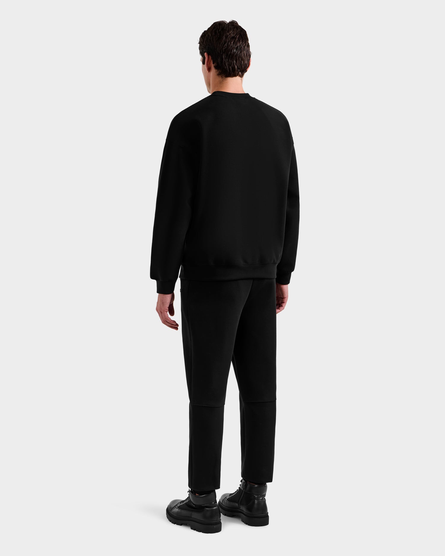 A man faces away from the camera, wearing the Rudsak DECLAN - 6525648 Black men’s crewneck sweatshirt with black pants and shoes against a plain light gray background.