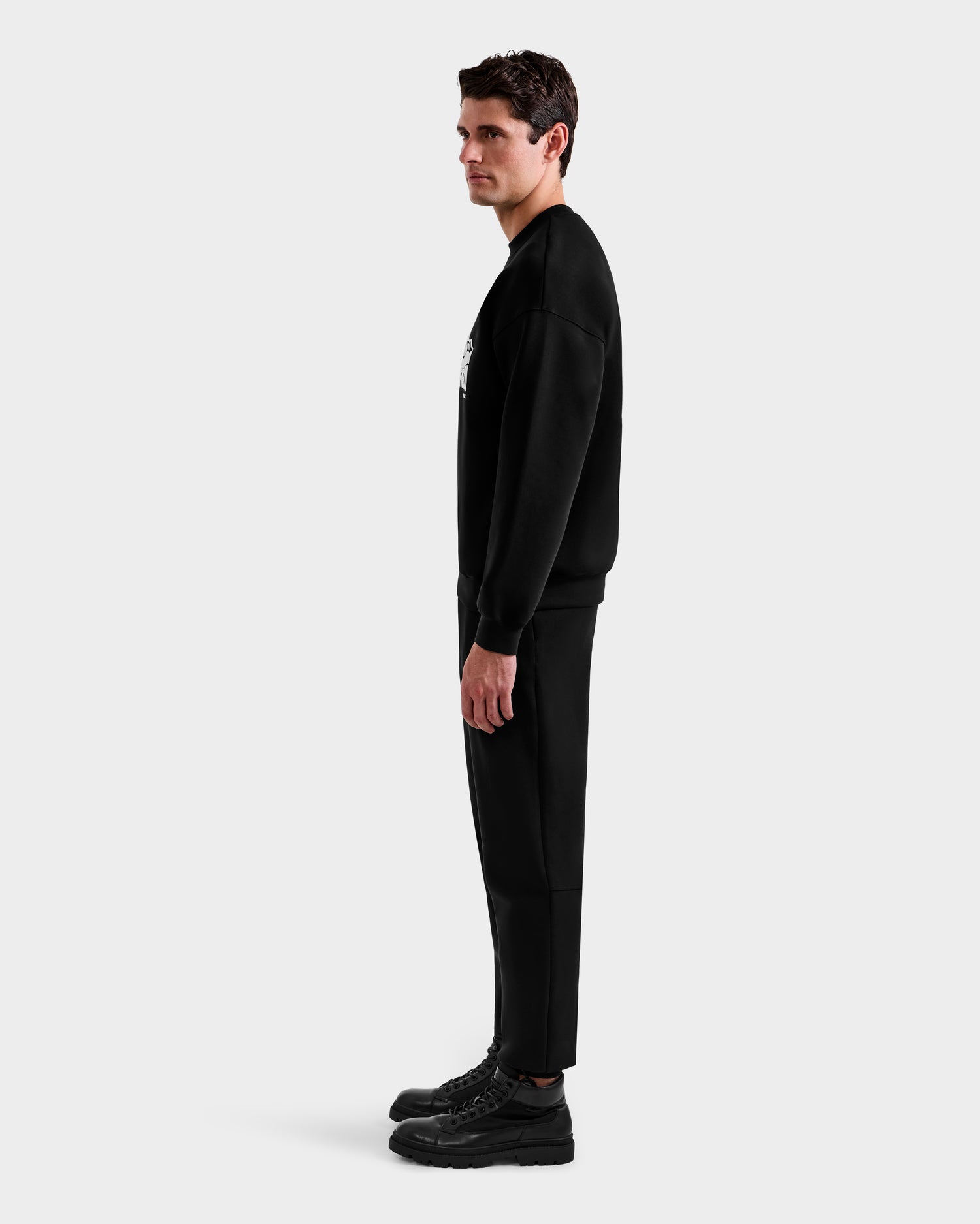 A man stands in profile against a plain background, wearing the Rudsak DECLAN - 6525648 Black men’s crewneck sweatshirt, paired with black pants and shoes, looking straight ahead with his arms relaxed by his sides.