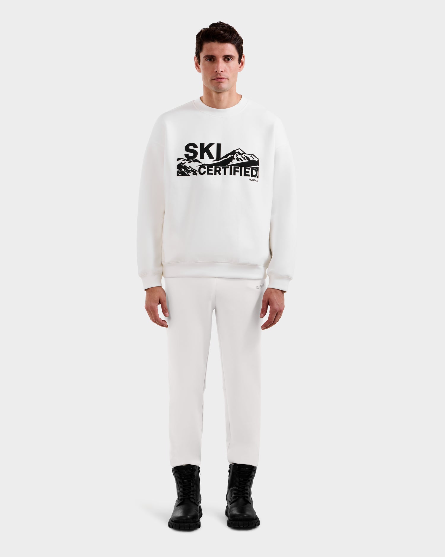 A man faces forward wearing the DECLAN - 6525648 Iceland white crewneck sweatshirt by Rudsak with SKI CERTIFIED and mountain graphics, styled with white pants and black boots against a plain light background.