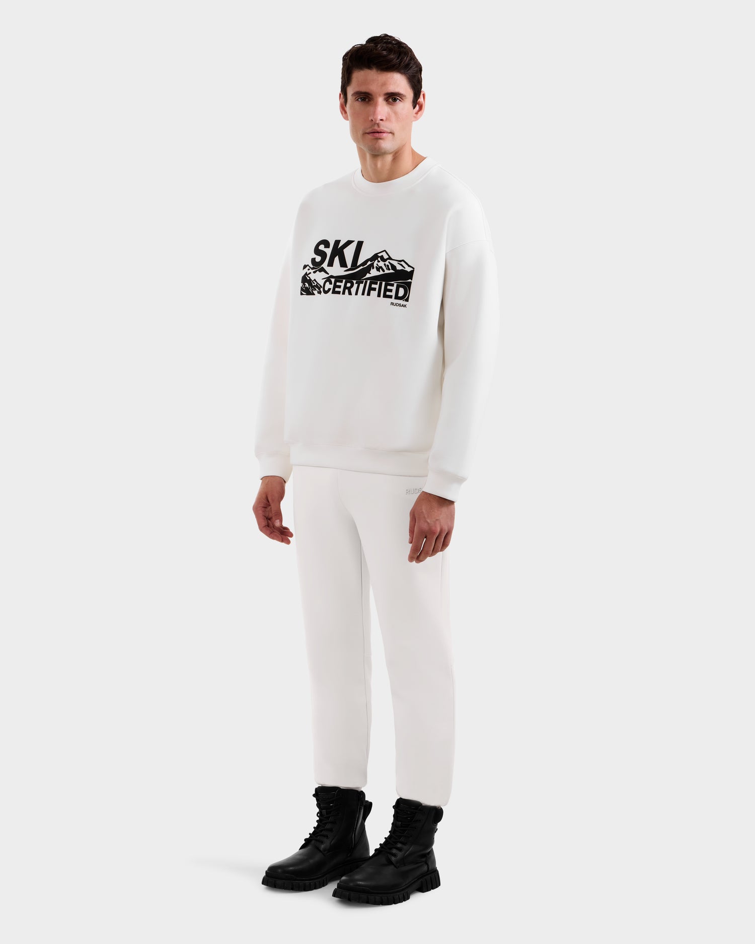 A man confidently poses against a white background wearing the Rudsak DECLAN - 6525648 Iceland men's crewneck sweatshirt featuring “SKI CERTIFIED” and mountain graphics, styled with white pants and black chunky boots.