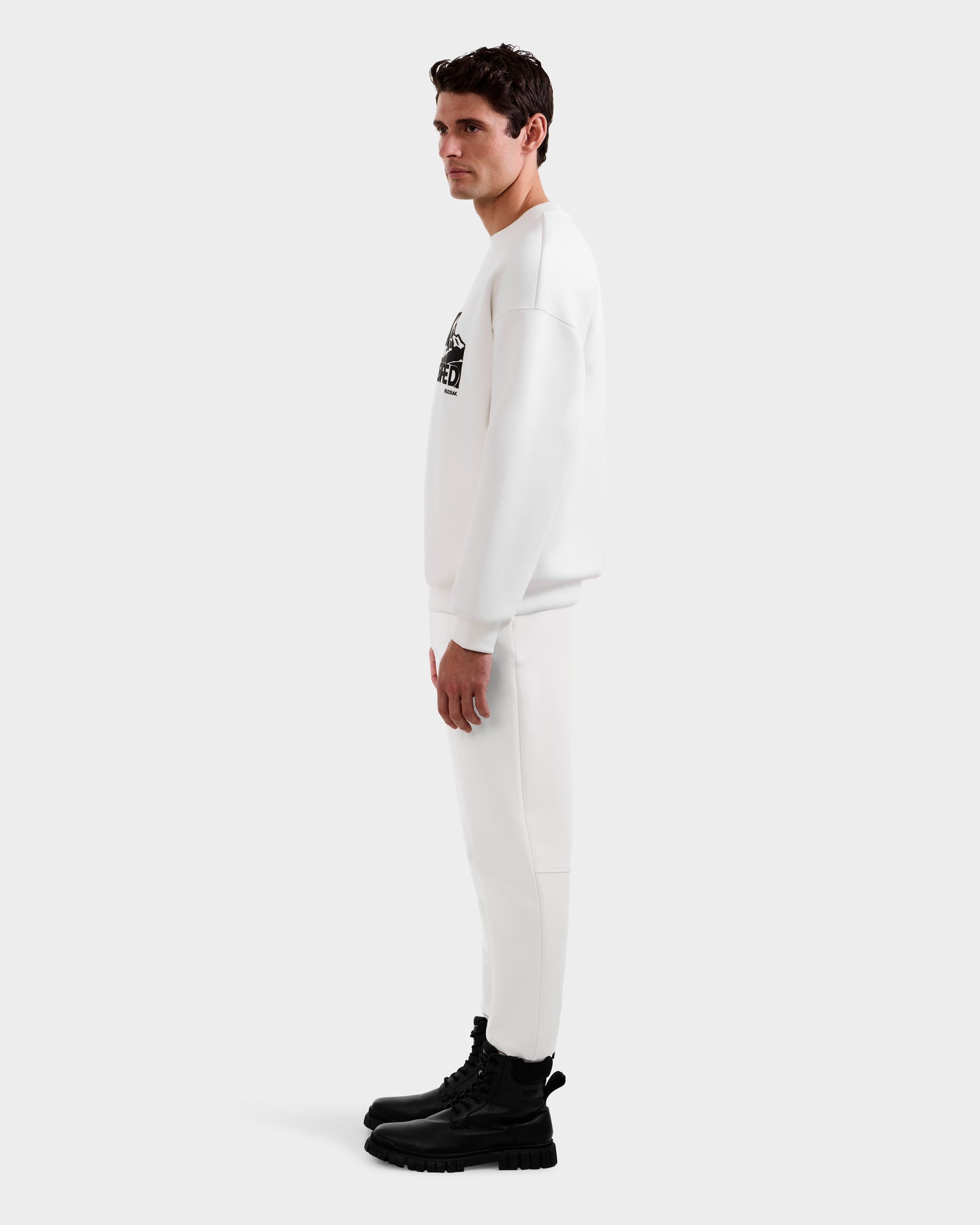 A man stands in profile wearing the Rudsak DECLAN - 6525648 Iceland men’s crewneck sweatshirt tracksuit with a chest graphic, paired with black combat boots, against a plain white background.