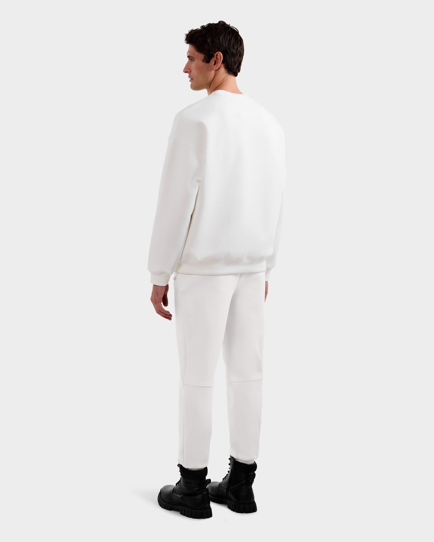 A man faces away from the camera in a Rudsak DECLAN - 6525648 Iceland men’s crewneck sweatshirt, paired with white pants and black boots, set against a plain light background.