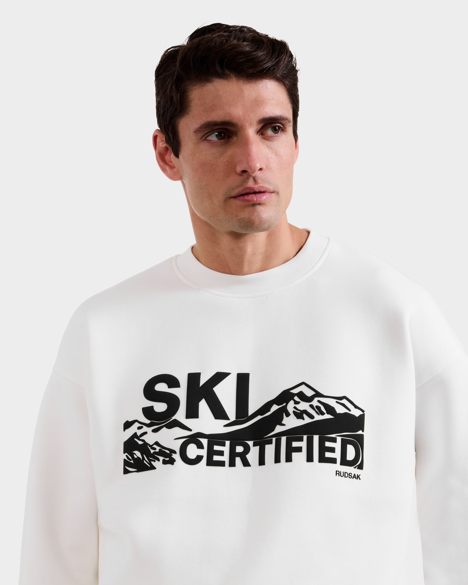 A man stands against a plain white background wearing the Rudsak DECLAN - 6525648 Iceland men's crewneck sweatshirt, featuring a black mountain graphic and the words "SKI CERTIFIED" on the front.