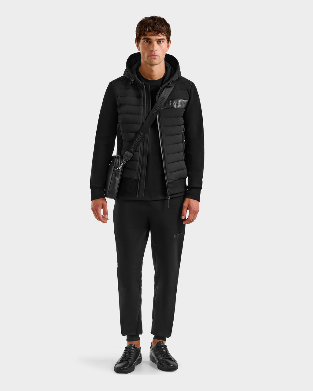A man with short curly brown hair wears the Rudsak DEE MP - 6125673 Black men’s lightweight down jacket, a black t-shirt, joggers, sneakers, and carries a black crossbody bag against a plain white background.