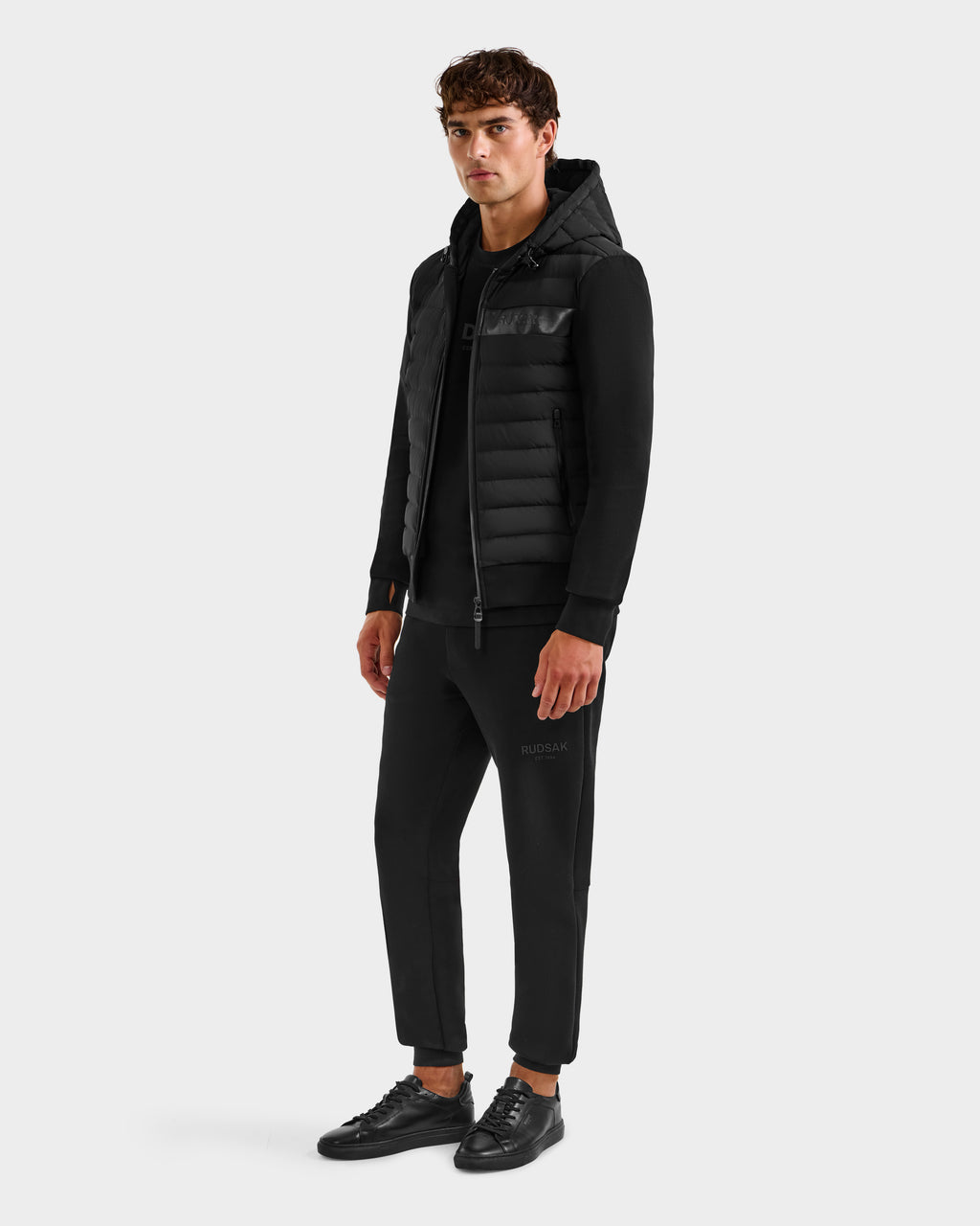 A man faces forward wearing the DEE MP - 6125673 Black lightweight down jacket by Rudsak, styled with black joggers and sneakers—a perfect transitional staple—set against a plain white background.