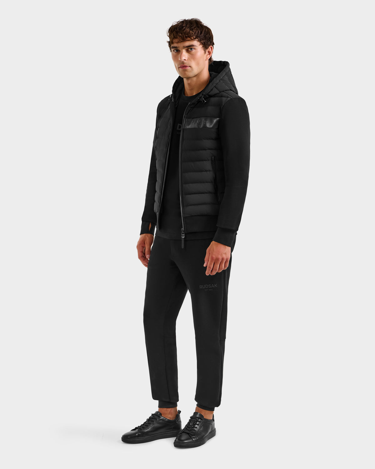 A man faces forward wearing the DEE MP - 6125673 Black lightweight down jacket by Rudsak, styled with black joggers and sneakers—a perfect transitional staple—set against a plain white background.