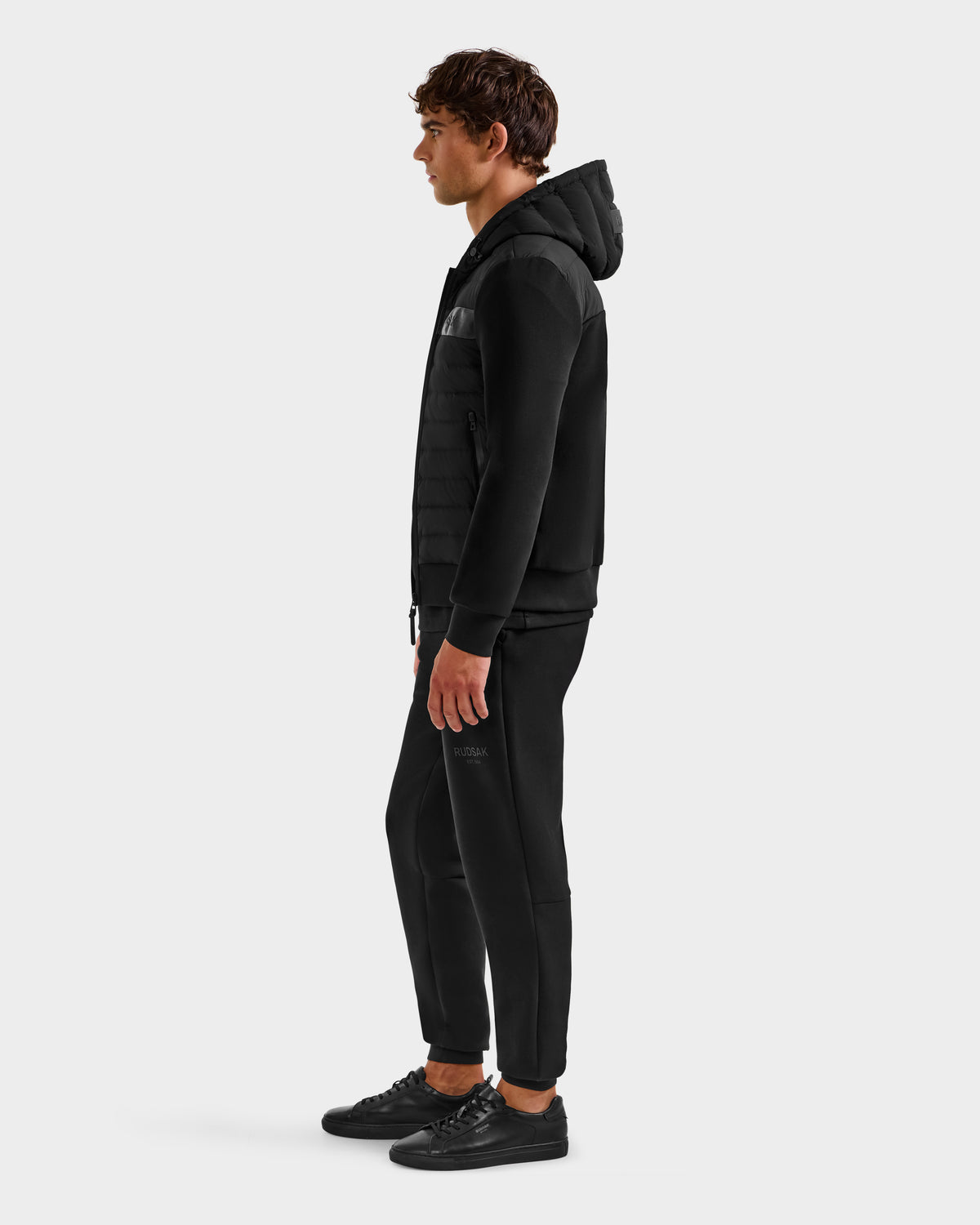 A man stands in profile against a plain background, wearing the Rudsak DEE MP - 6125673 Black men's lightweight down jacket with a quilted front—filled with RDS-certified duck down—ideal as a transitional staple.