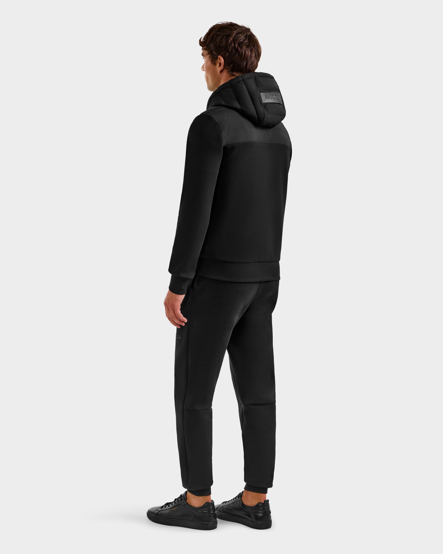 Facing away, a person wears the Rudsak DEE MP - 6125673 Black men's lightweight down jacket with black jogger pants and sneakers against a plain light gray background.