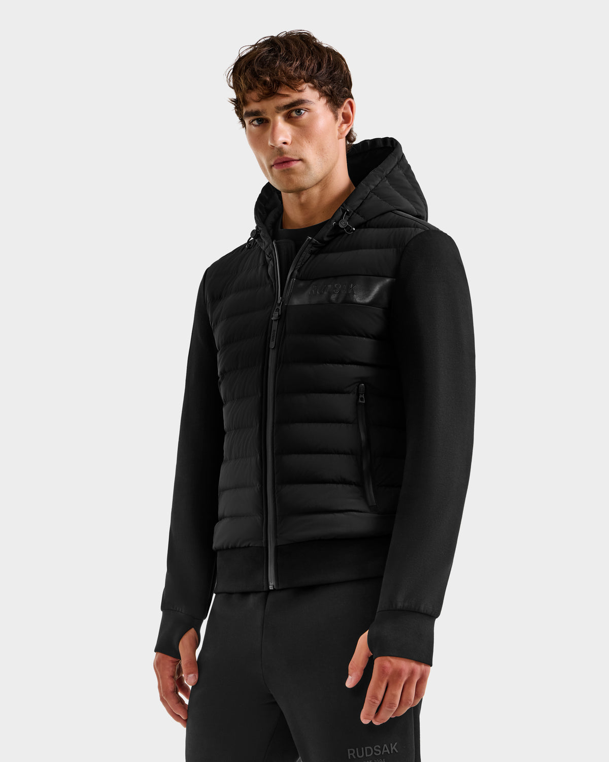 A young man stands against a plain light gray background, wearing the Rudsak DEE MP - 6125673 Black men’s lightweight down jacket with zippered pockets and black pants, looking directly at the camera with a neutral expression.