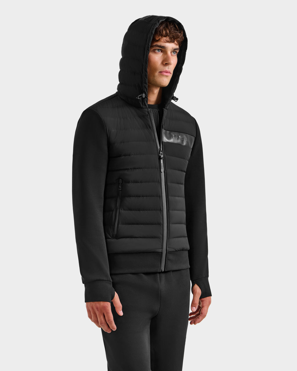 A young man wears the Rudsak DEE MP - 6125673 Black men’s lightweight down jacket, filled with RDS-certified duck down, paired with matching black pants and stands against a plain light gray background, facing slightly right.