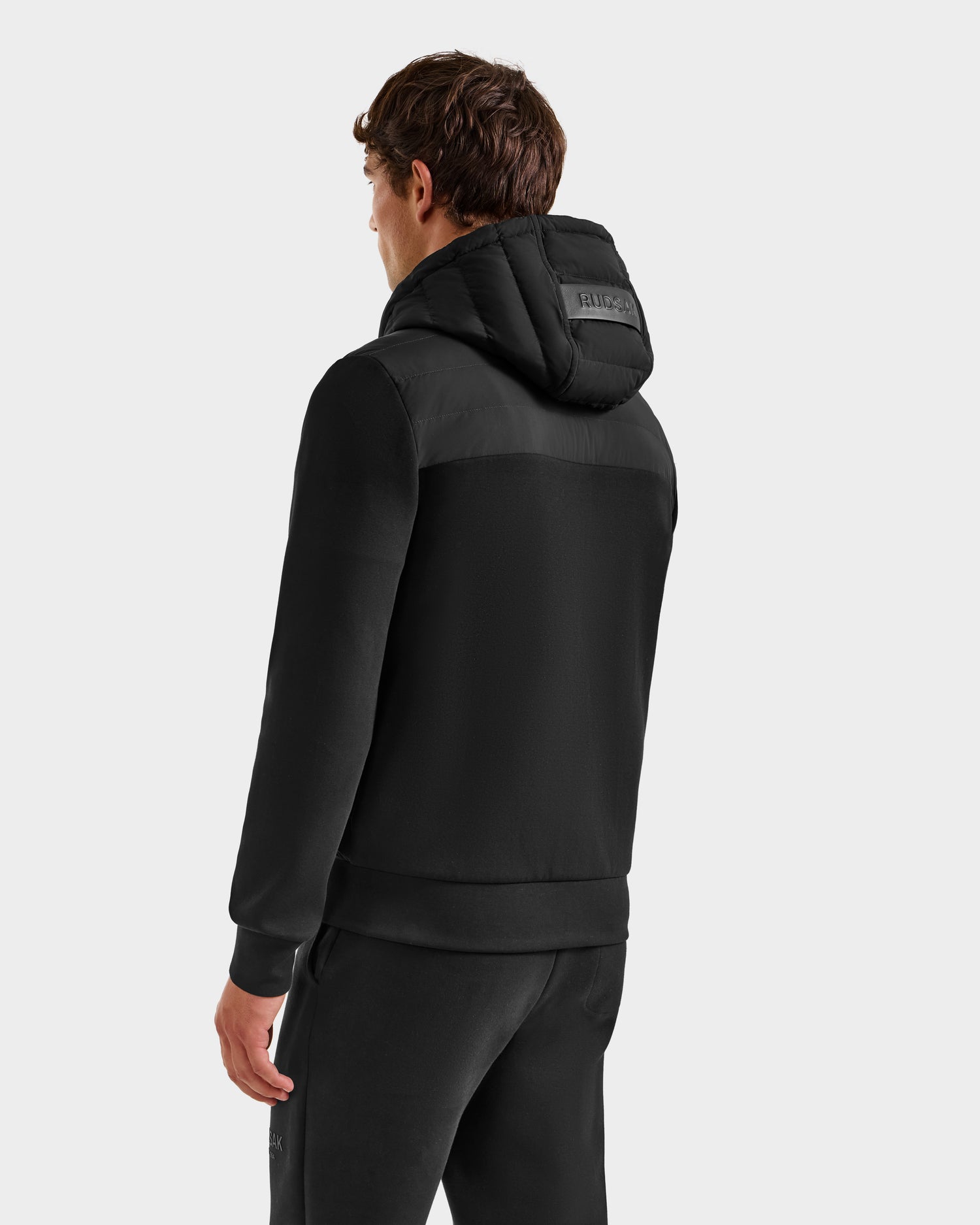 A person is shown from the back wearing the Rudsak DEE MP - 6125673 Black men's lightweight down jacket and black pants, standing against a plain light gray background.