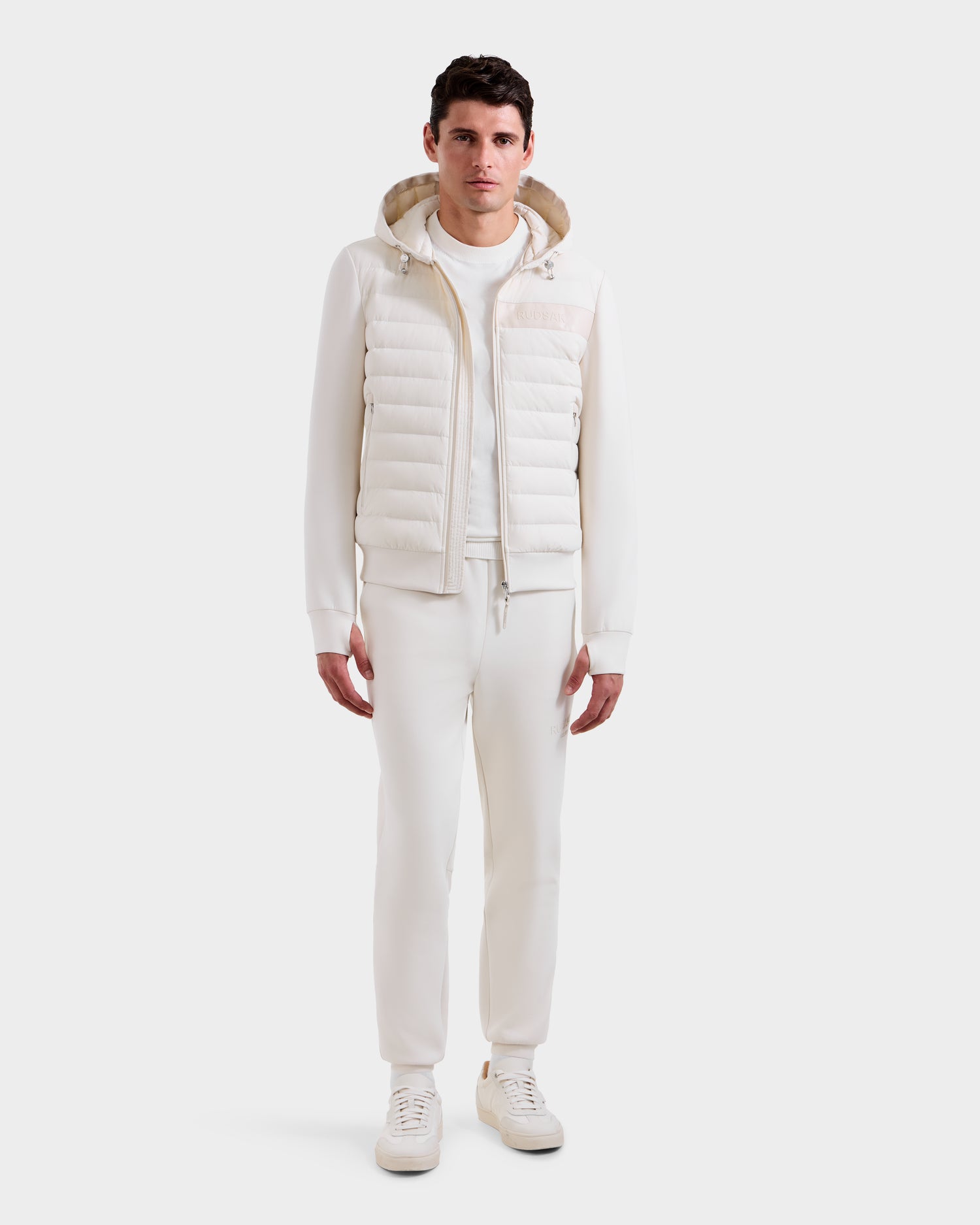 A man faces forward in an all-white outfit, featuring the Rudsak DEE MP - 6125673 Vanilla men’s lightweight down jacket with RDS-certified duck down, paired with a sweatshirt, pants, and sneakers against a plain white background.