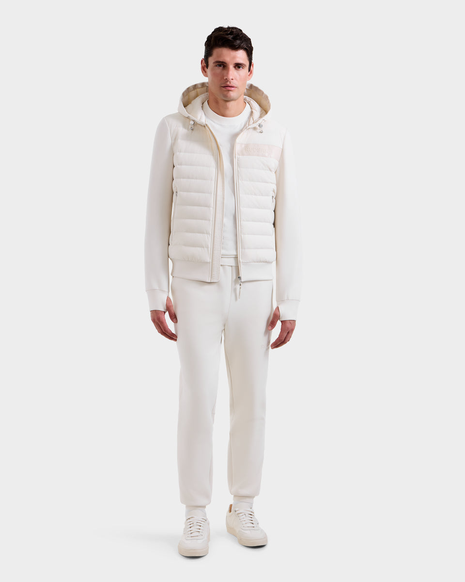 A man faces forward in an all-white outfit, featuring the Rudsak DEE MP - 6125673 Vanilla men’s lightweight down jacket with RDS-certified duck down, paired with a sweatshirt, pants, and sneakers against a plain white background.