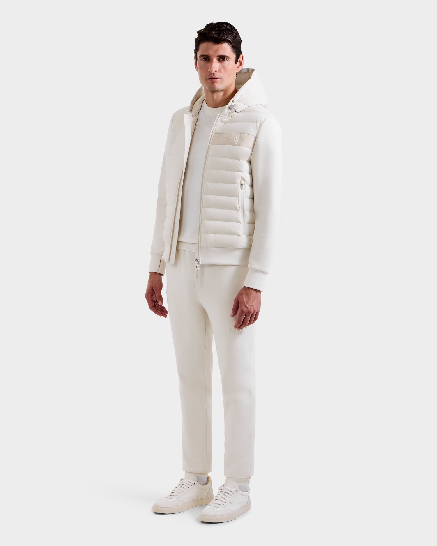 A man faces forward in an all-white outfit, featuring the Rudsak DEE MP - 6125673 Vanilla men’s lightweight down jacket with quilted hood, sweatshirt, pants, and sneakers against a plain white background—a perfect transitional staple.