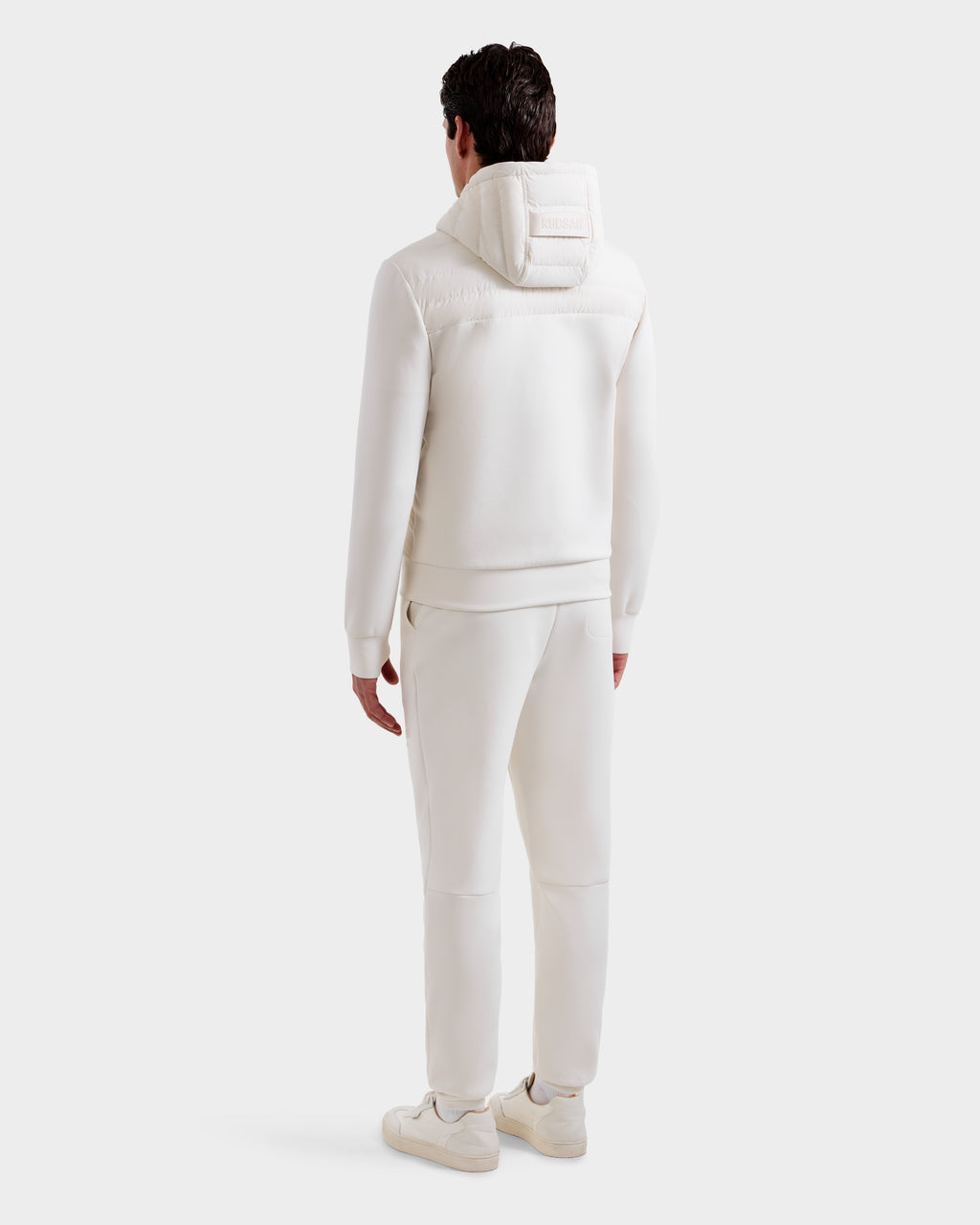 A man stands facing away in the Rudsak DEE MP - 6125673 Vanilla hooded tracksuit, paired with matching pants and sneakers, against a plain light background—an ideal transitional staple.