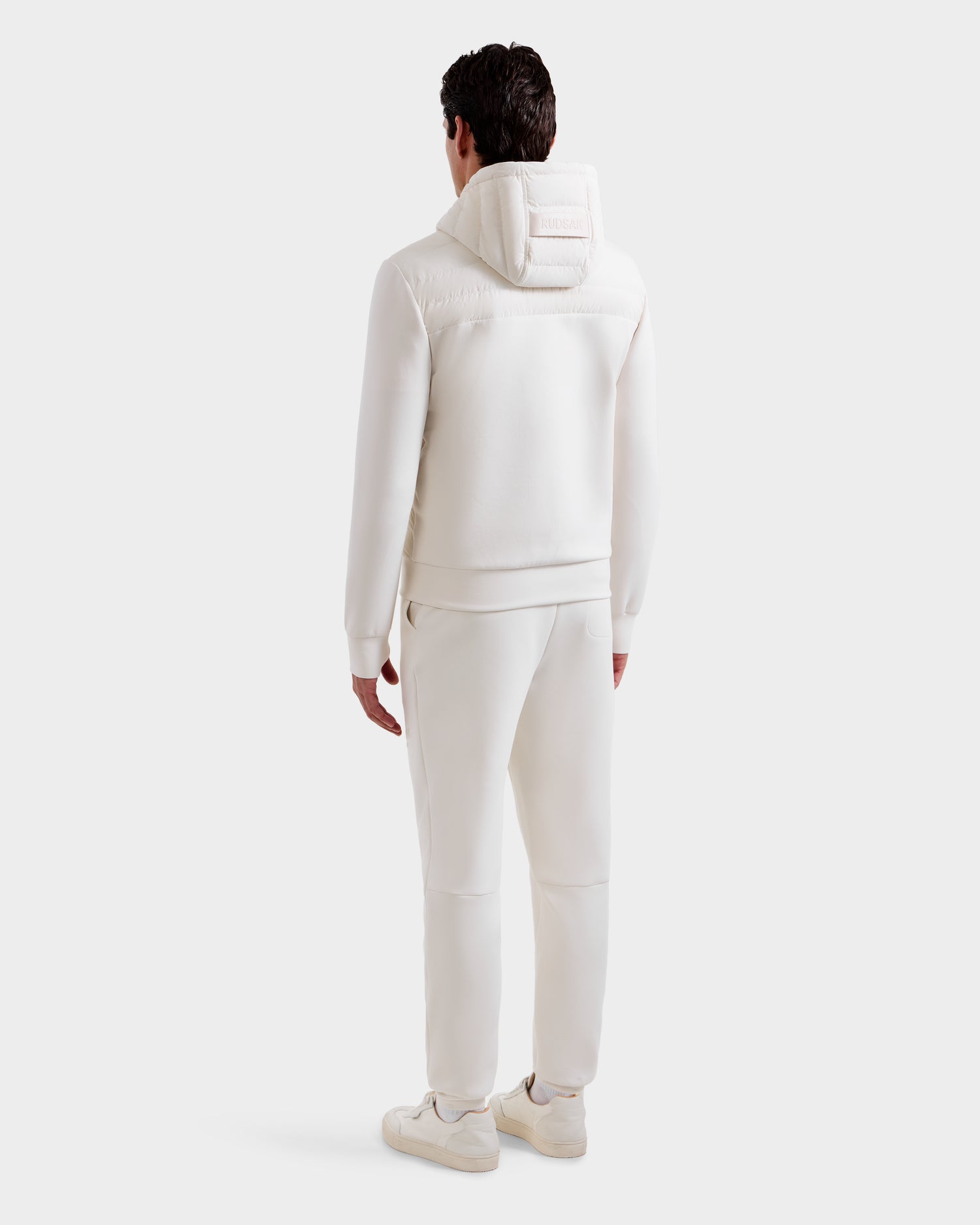 A man stands facing away in the Rudsak DEE MP - 6125673 Vanilla hooded tracksuit, paired with matching pants and sneakers, against a plain light background—an ideal transitional staple.