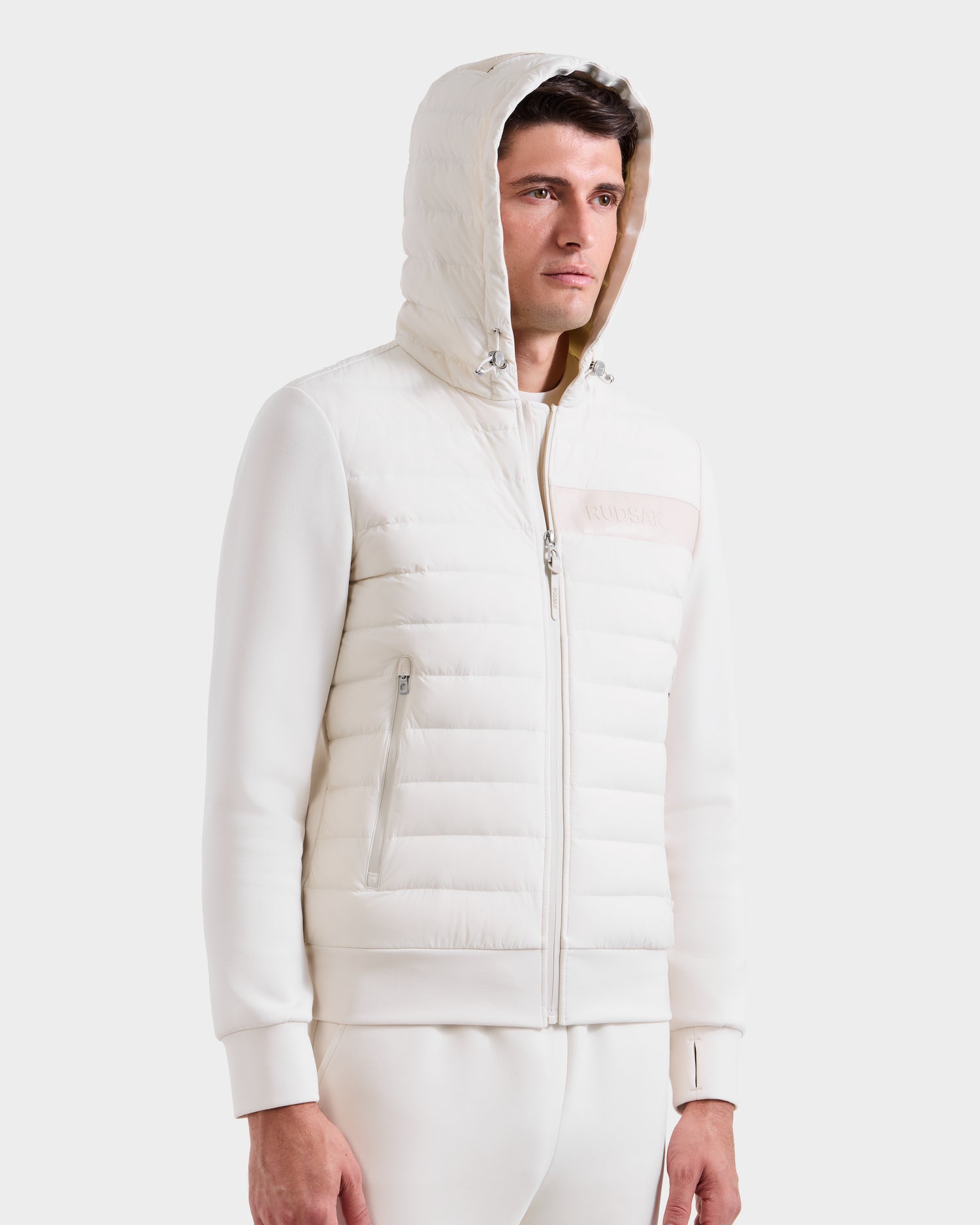 A man wears the Rudsak DEE MP - 6125673 Vanilla, a white lightweight down jacket with quilted panels and smooth sleeves, standing against a plain light background—the perfect transitional staple.