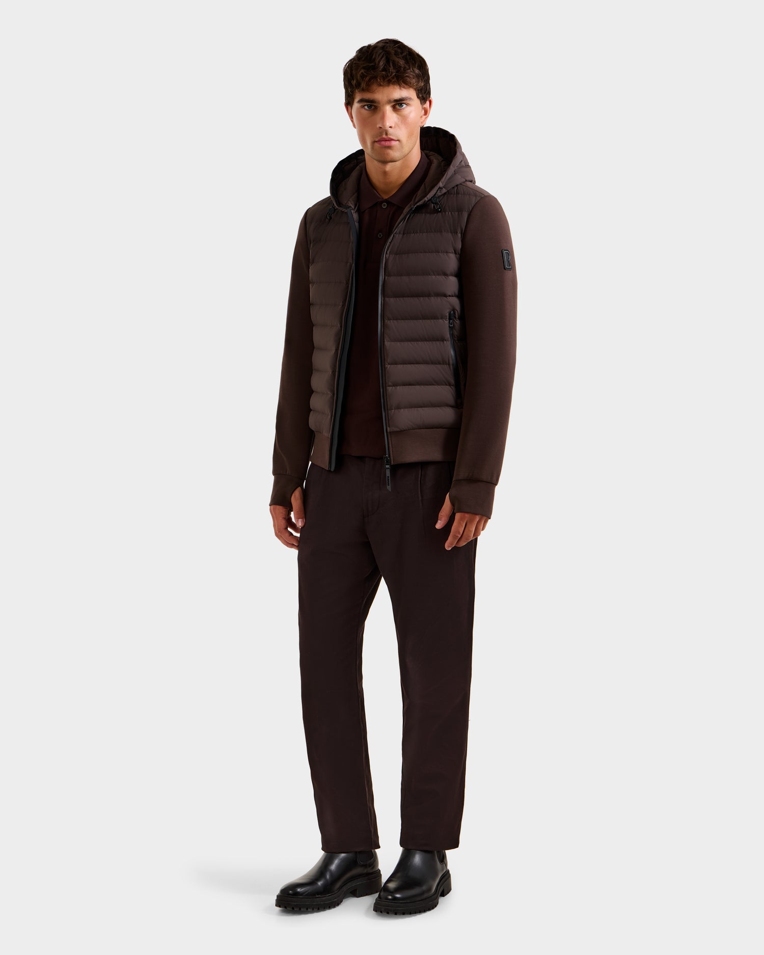 A man with short dark hair and a neutral expression wears the Rudsak DEE MAT - 6124028 Espresso men's lightweight hybrid down jacket, paired with matching brown pants, a dark polo, and black boots for an understated urban look.