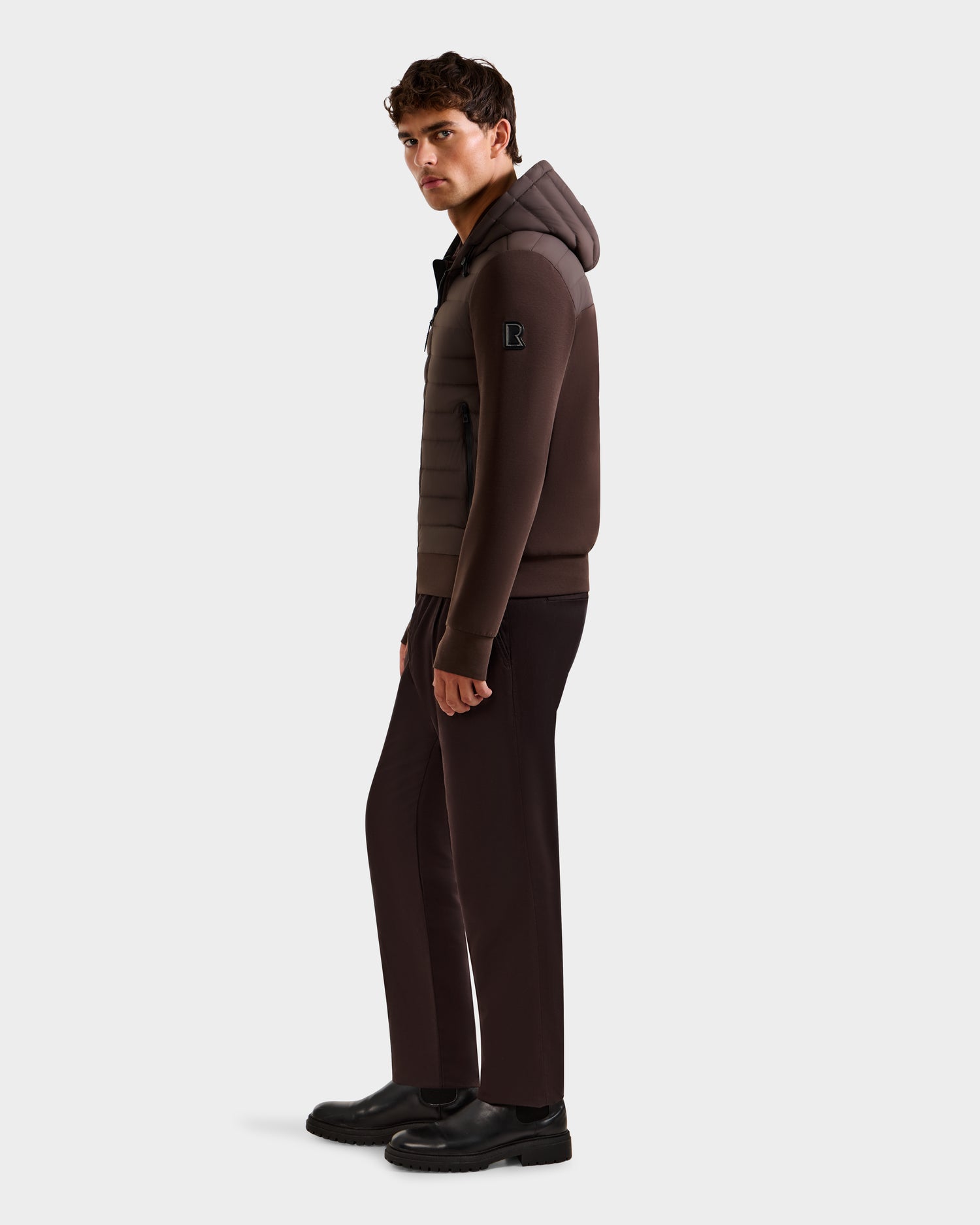 A man stands sideways, looking forward, dressed in the Rudsak DEE MAT - 6124028 Espresso jacket with matching pants and black shoes against a plain white background.