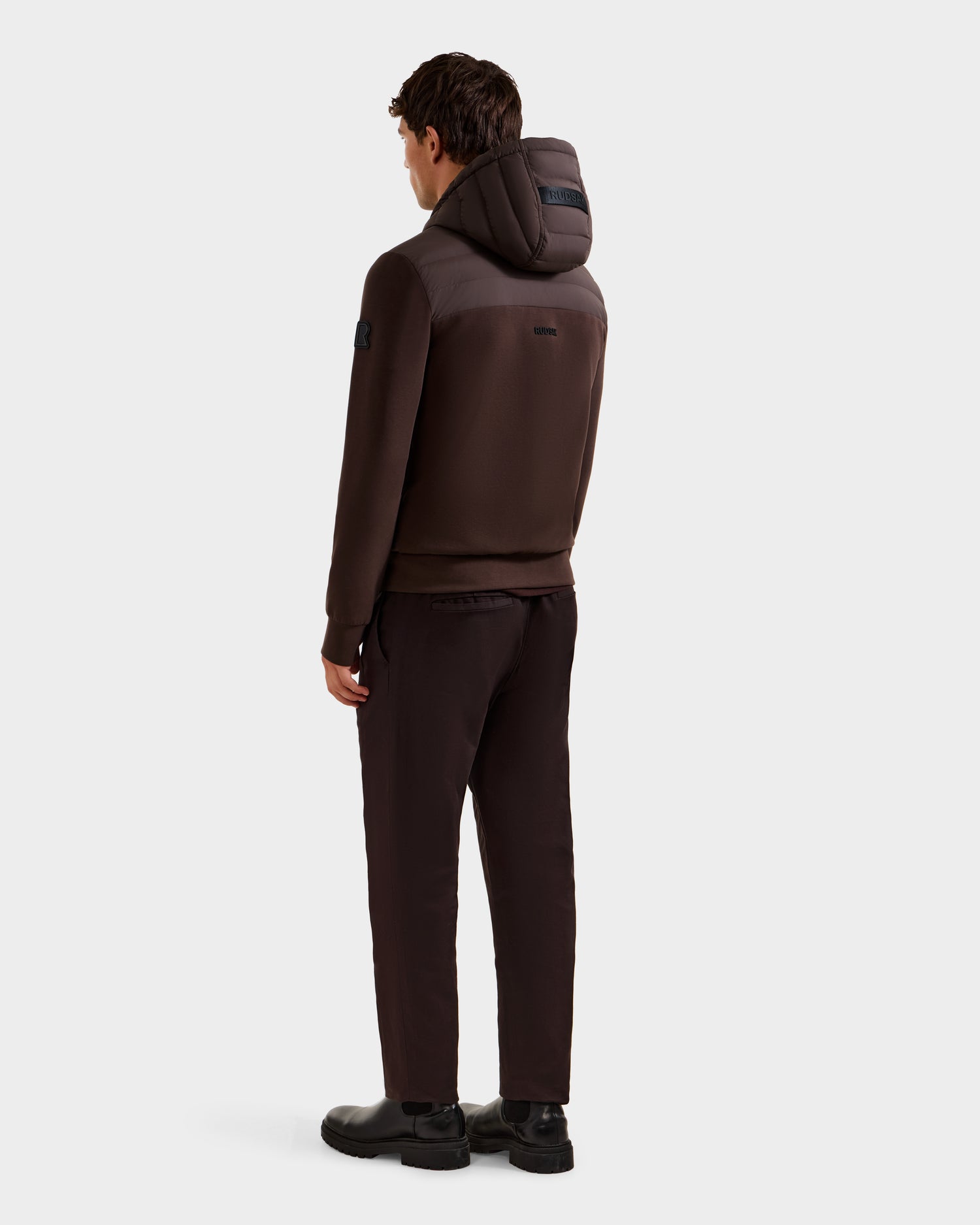 A man, facing away, wears the Rudsak DEE MAT - 6124028 Espresso jacket and matching pants with black shoes against a plain light background.