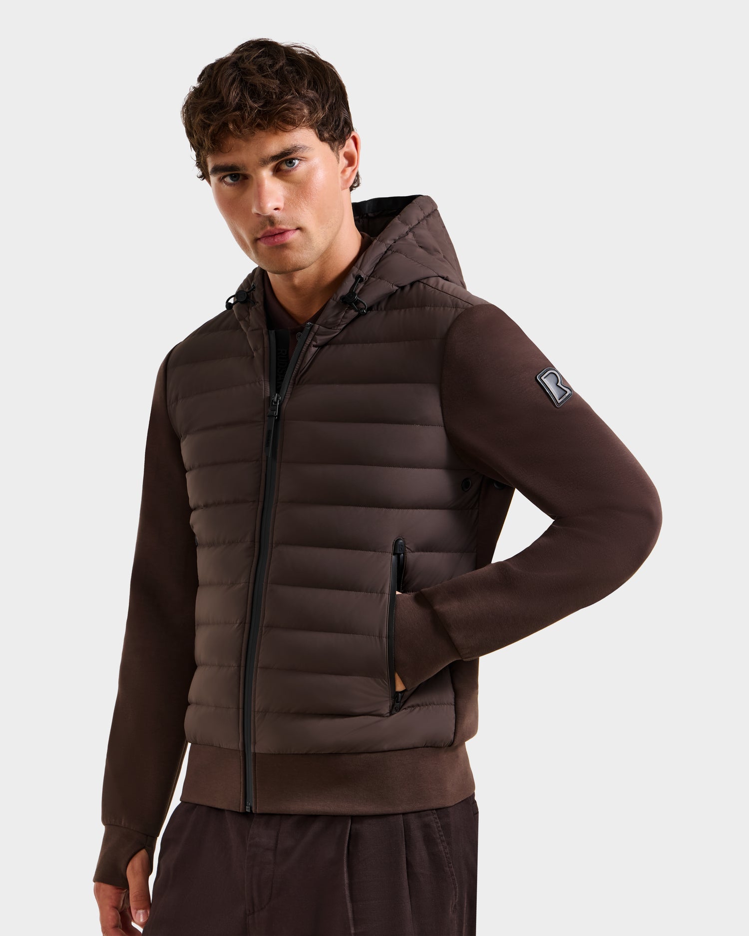 A man wears the Rudsak DEE MAT - 6124028 Espresso men’s lightweight hybrid down jacket with zippered pockets and matching brown pants, standing against a plain light background and looking at the camera with a neutral expression.